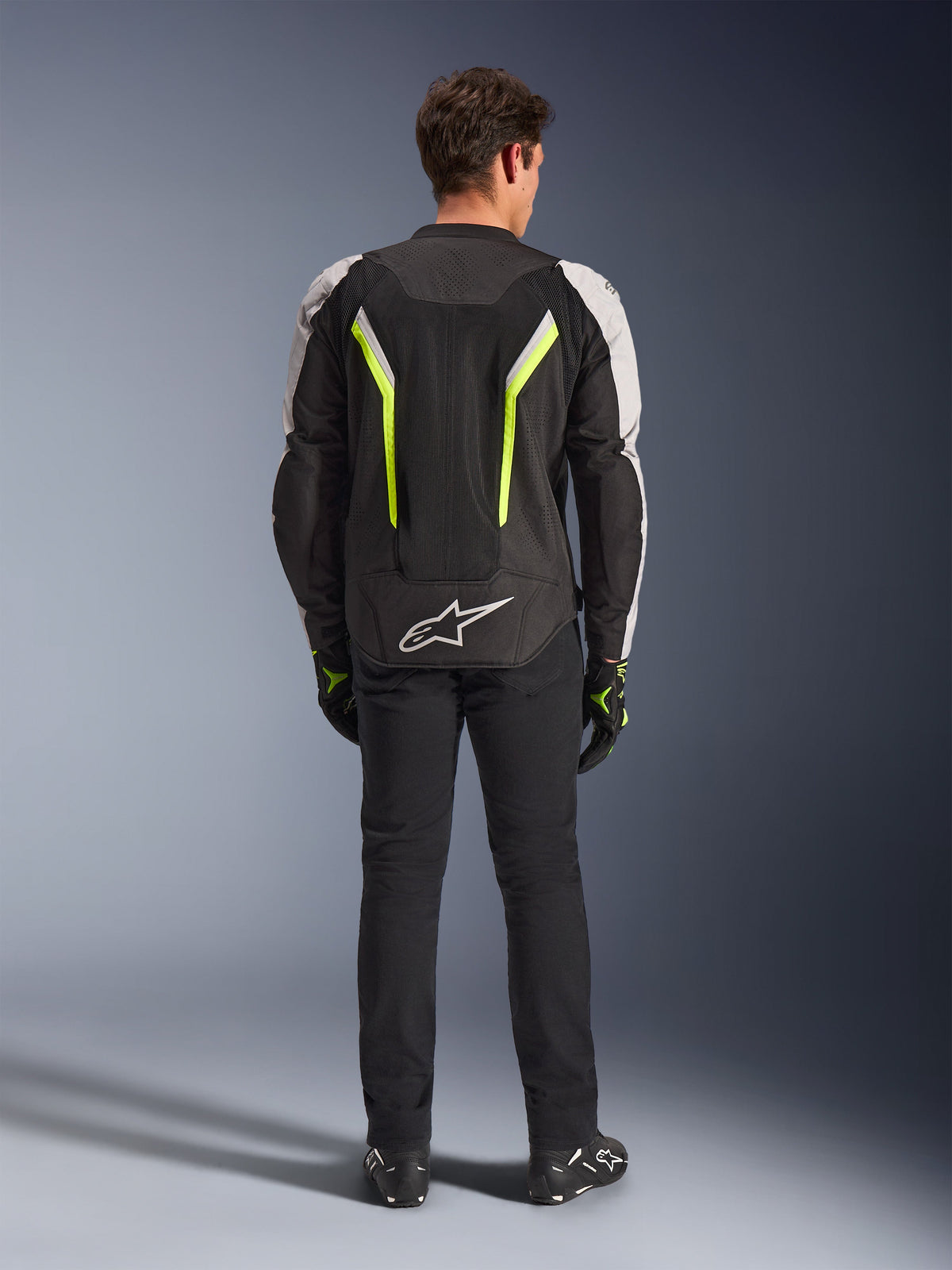 Alpinestars AST V3 Air Jacket, giacca da moto in tessuto, Nero, Argento e Giallo Fluo, vista posteriore che mostra i pannelli di ventilazione in rete e gli accenti fluorescenti, indossata con pantaloni neri e guanti da moto, abbigliamento tecnico sportivo