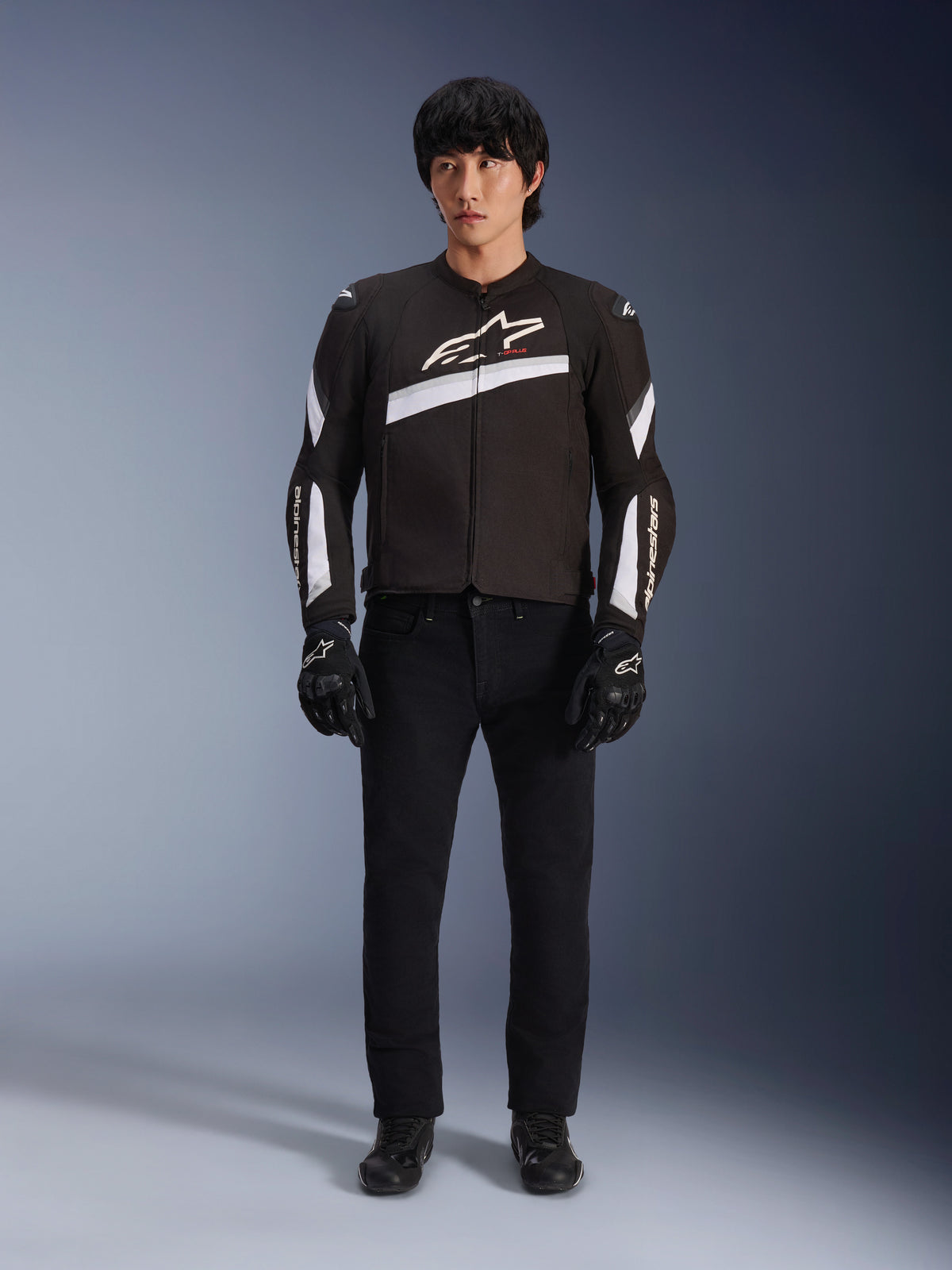 Giacca Alpinestars T-GP Plus R V4, nero e bianco, giacca da moto sportiva in tessuto, indossata da un modello con pantaloni e guanti neri, con vistoso logo Alpinestars bianco su petto e maniche, design elegante di ispirazione racing