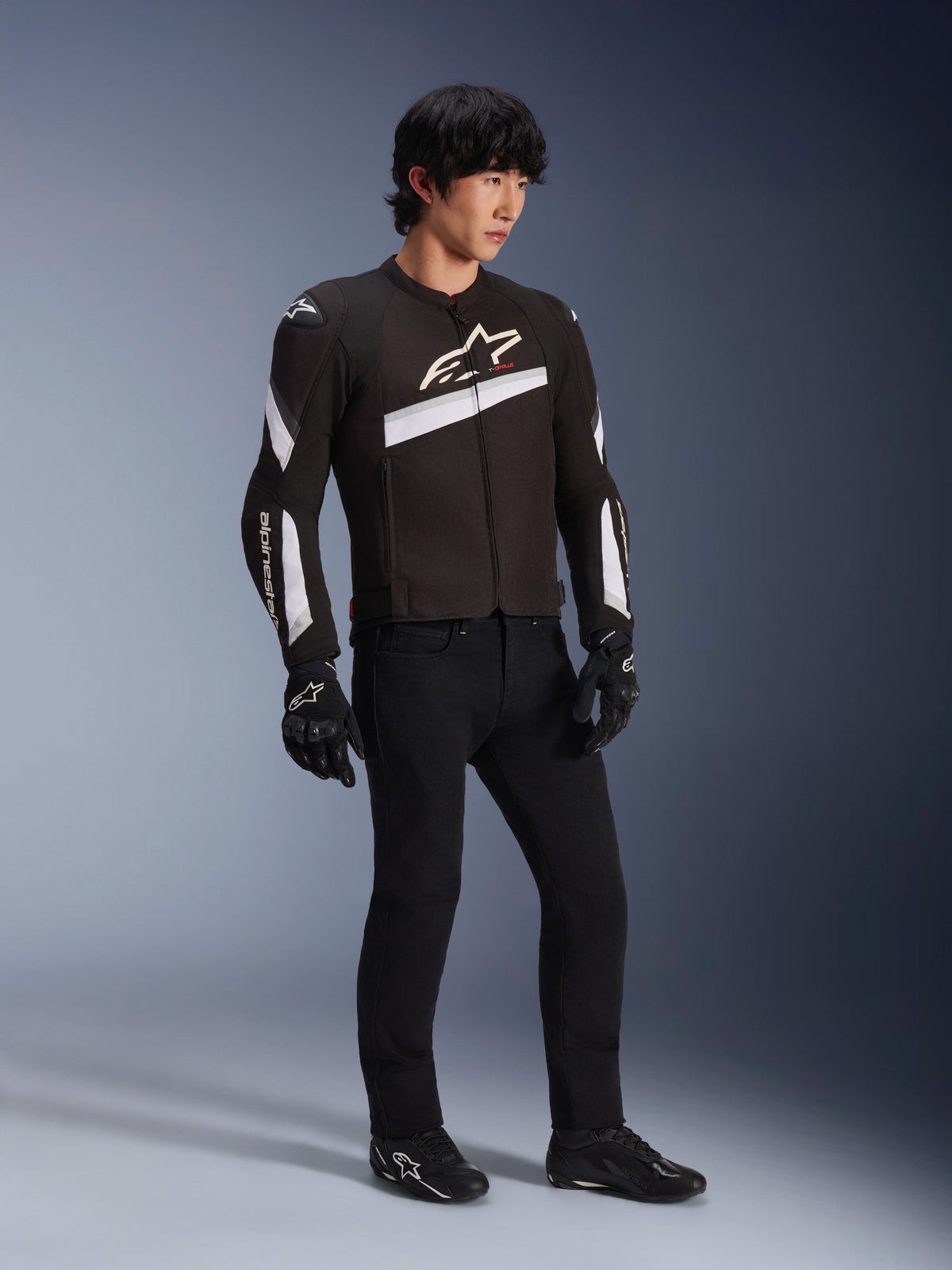 Giacca Alpinestars T-GP Plus R V4, giacca da moto in tessuto nera e bianca indossata da un modello con pantaloni da guida e guanti neri, con loghi Alpinestars bianchi sul petto e sulle maniche, design sportivo per prestazioni su strada