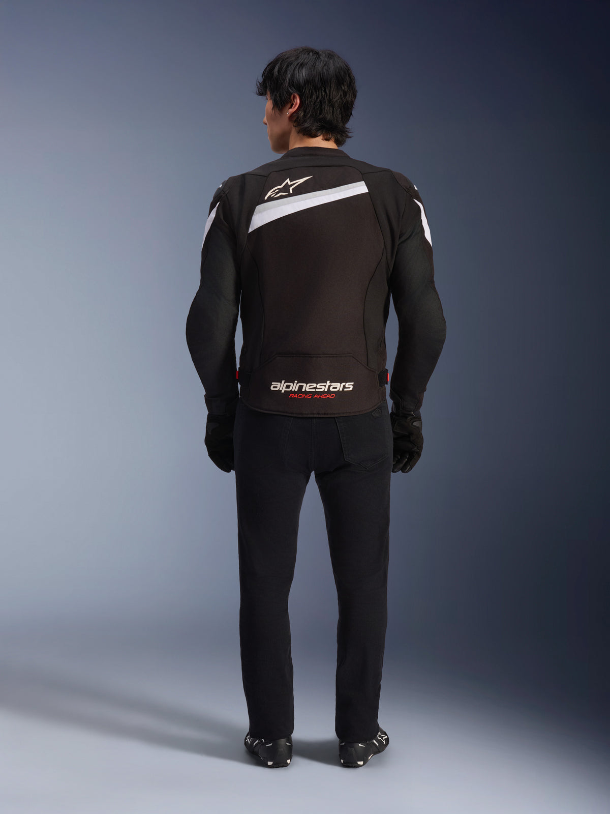 Giacca Alpinestars T-GP Plus R V4, giacca da moto sportiva in tessuto nero e bianco, vista posteriore con logo Alpinestars bianco e marchio 'RACING AHEAD', indossata con pantaloni e stivali neri, design aerodinamico per prestazioni elevate.