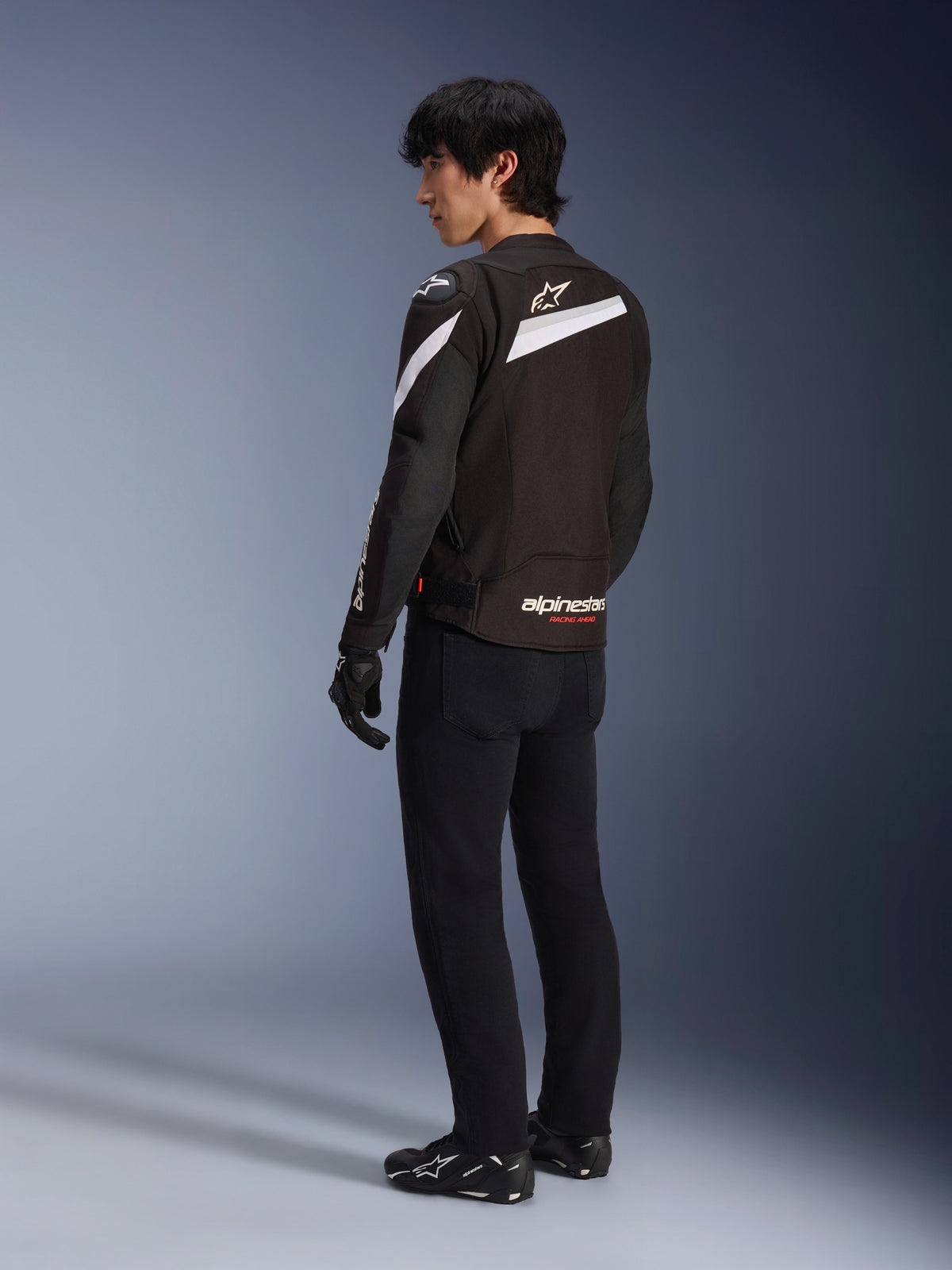 Pilota uomo che indossa la giacca Alpinestars T-GP Plus R V4 in nero e bianco, vista posteriore che mostra accenti diagonali bianchi e logo sulla parte bassa della schiena, abbinata a pantaloni da moto neri, guanti e stivali tecnici, abbigliamento da moto in tessuto con vestibilità sportiva