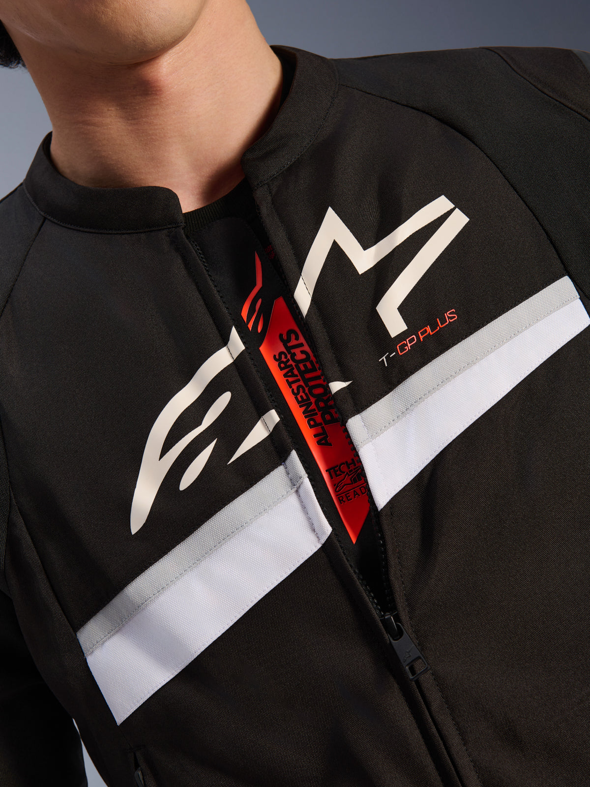 Giacca Alpinestars T-GP Plus R V4, primo piano di petto e collo, tessuto nero con logo a stella bianco e marchio T-GP PLUS, striscia orizzontale bianca e grigia, cerniera aperta che rivela un'etichetta interna rossa con scritta Tech-Air Ready