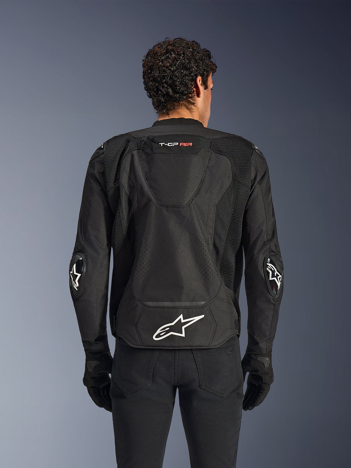 Alpinestars T-GP Air Jacket, nera, vista posteriore di una giacca da moto in tessuto, caratterizzata da gobba aerodinamica, ampi pannelli di ventilazione in rete e logo Alpinestars bianco sulla parte inferiore della schiena, abbinata a guanti e pantaloni da moto neri