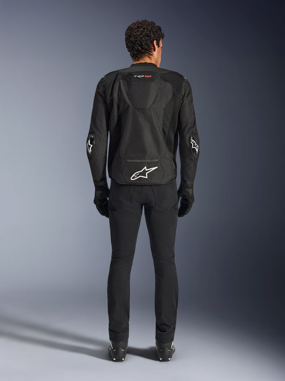 Alpinestars T-GP Air Jacket, giacca da moto in tessuto, nera, vista posteriore di un uomo con pannelli di ventilazione in rete, una gobba aerodinamica e loghi Alpinestars bianchi sulla parte inferiore della schiena e sui gomiti, indossata con pantaloni da guida e guanti neri