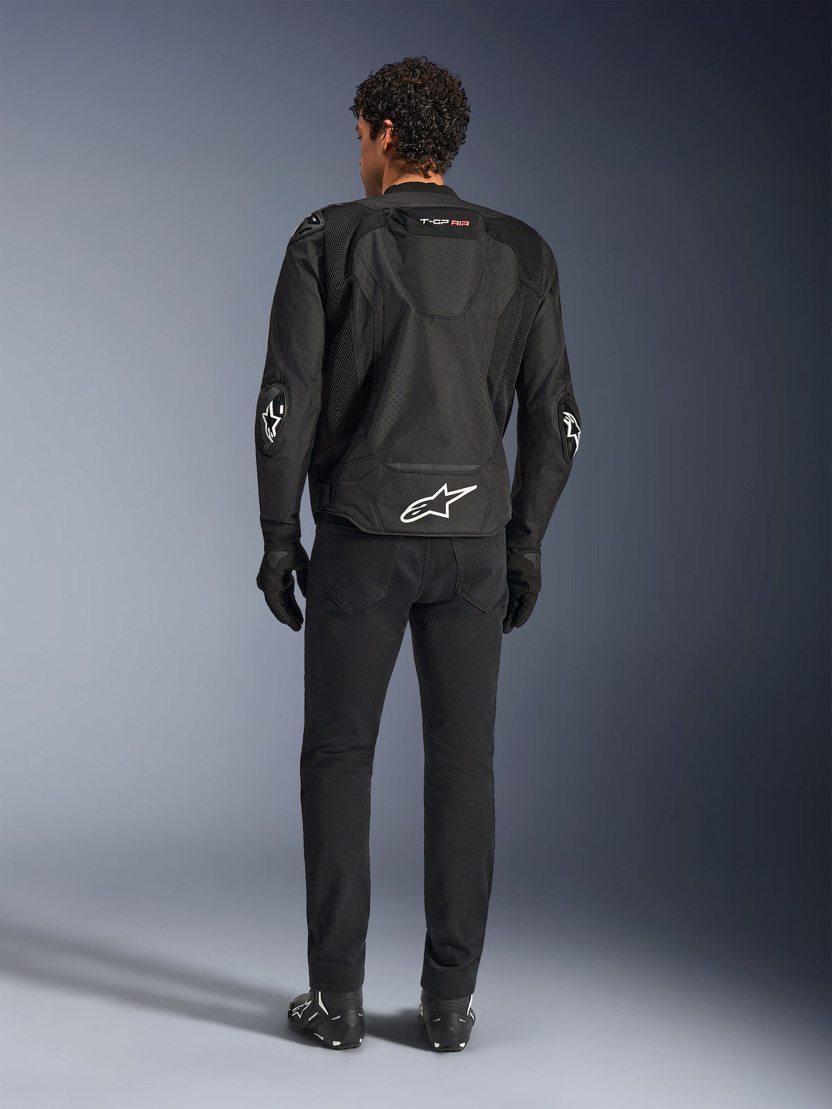 Giacca Alpinestars T-GP Air, nero, vista posteriore su modello, che mostra i pannelli di ventilazione in rete 3D e la gobba aerodinamica, con logo a stella bianco sulla parte bassa della schiena e sui gomiti, indossata con pantaloni e guanti da moto neri, abbigliamento tecnico sportivo.
