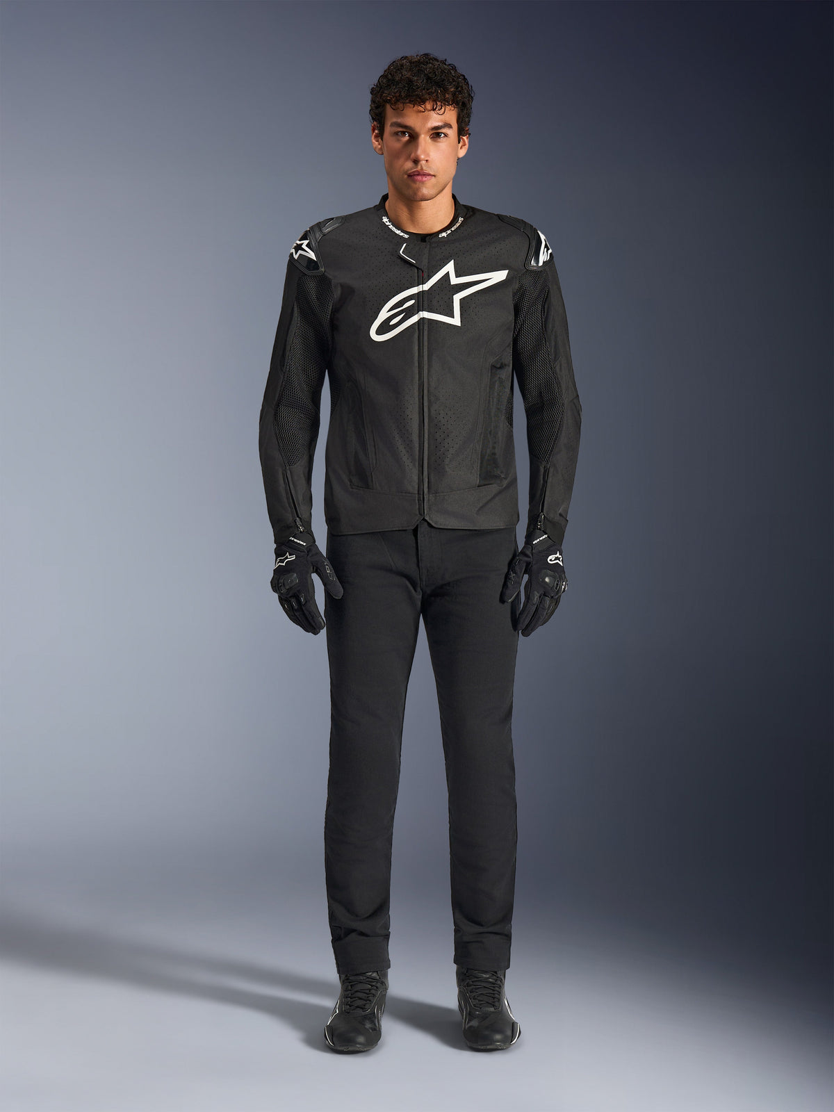 Alpinestars T-GP Air Jacket, giacca da moto in tessuto nero con un grande logo Astars bianco sul petto, indossata da un modello con pantaloni da guida e guanti neri, con pannelli traforati e ventilazione in rete per la guida sportiva