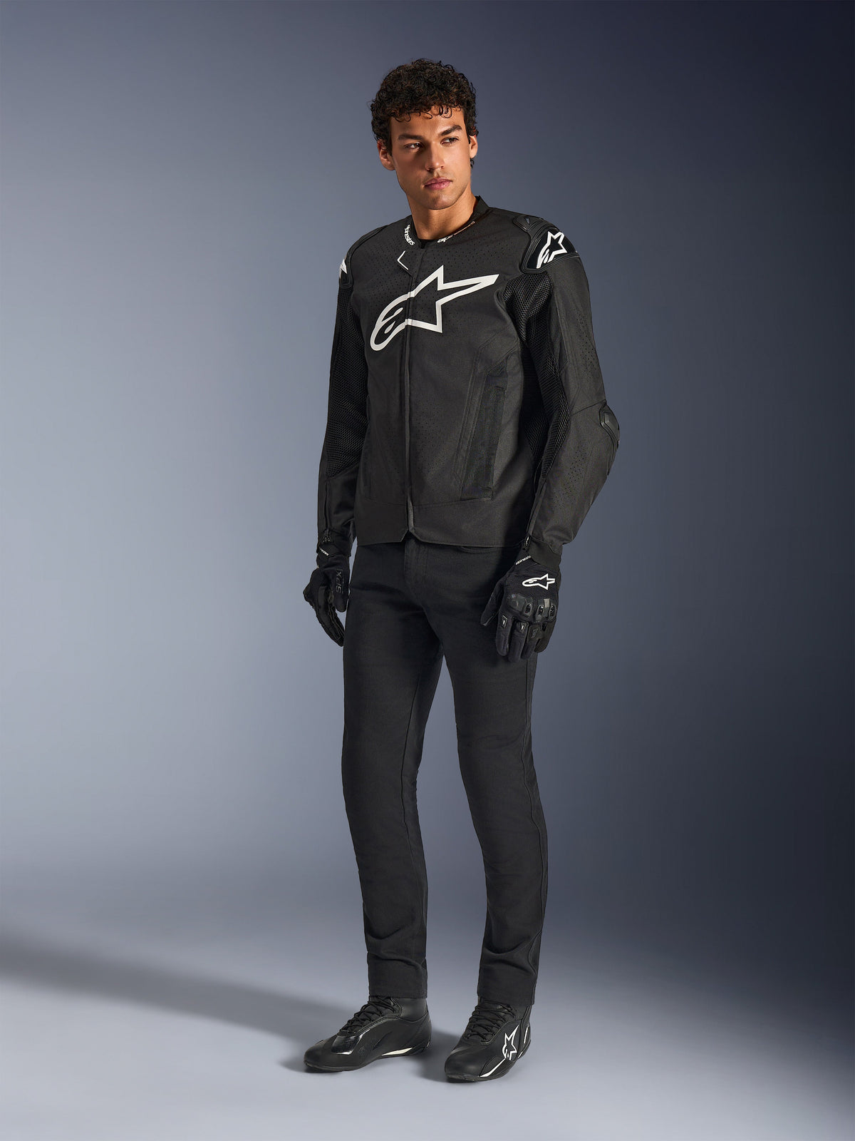 Alpinestars T-GP Air Jacket, giacca da moto in tessuto nero, indossata da un modello con pantaloni e guanti neri coordinati, con un evidente logo Alpinestars bianco sul petto e sulle spalle, pannelli in mesh ventilati e una vestibilità sportiva di derivazione racing