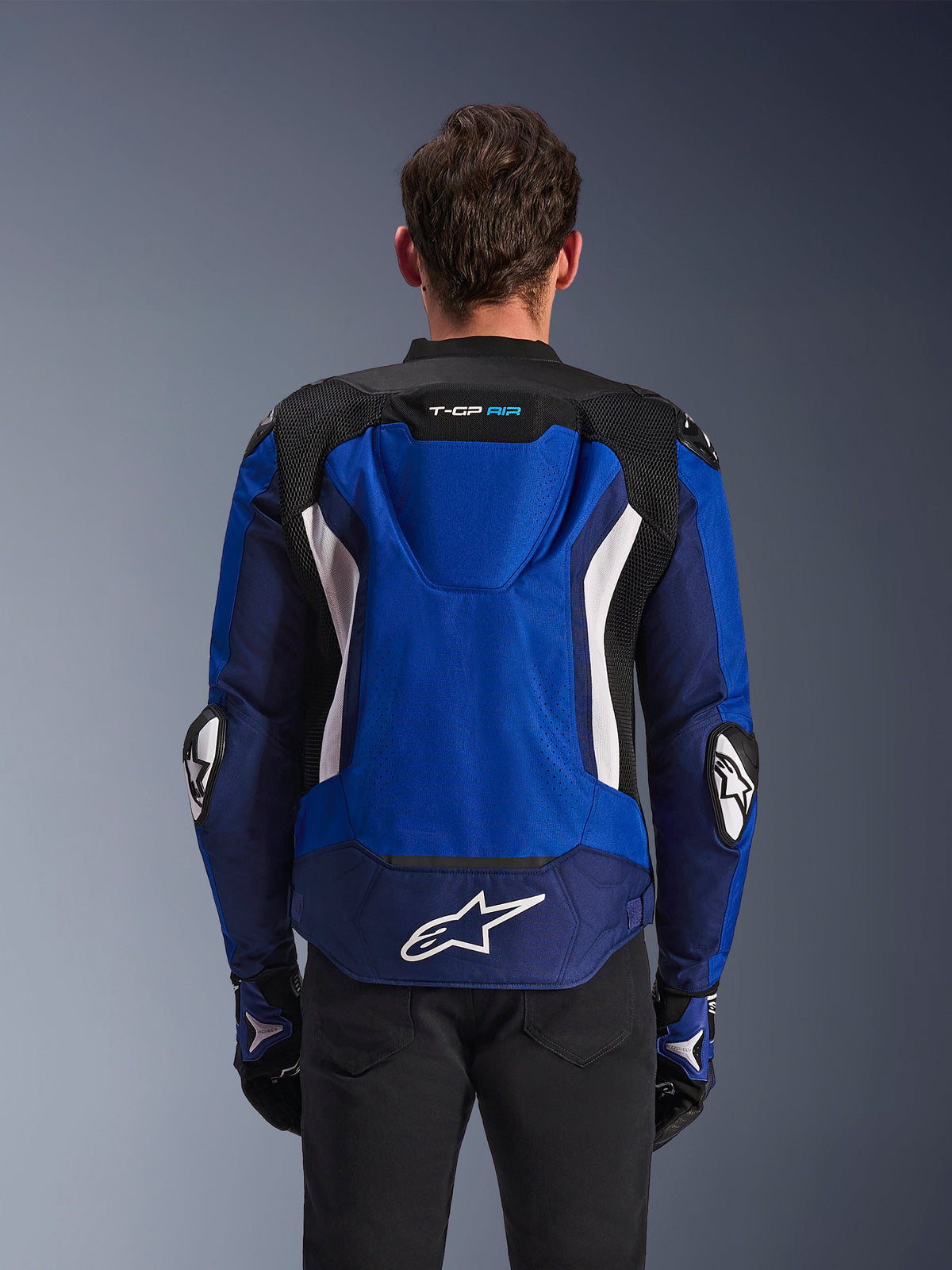 Giacca Alpinestars T-GP Air, giacca da moto sportiva in tessuto, blu navy brillante e bianco, vista posteriore con gobba aerodinamica integrata e grande logo bianco sulla parte bassa della schiena, indossata con guanti da moto coordinati blu e neri