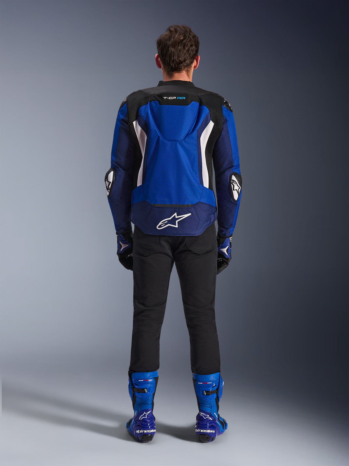 Giacca Alpinestars T-GP Air, Bright Blue Navy White, vista posteriore con gobba aerodinamica e pannelli di ventilazione in mesh, logo Alpinestars bianco sulla parte inferiore della schiena, indossata con pantaloni neri e stivali racing blu