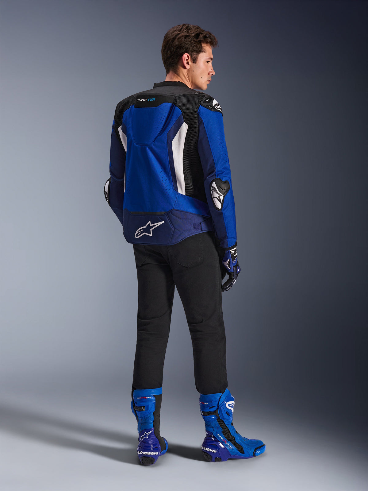 Alpinestars Giacca T-GP Air, giacca sportiva in tessuto, blu brillante, navy e bianco, vista posteriore su un modello con pantaloni neri e stivali blu e bianchi coordinati, con logo Alpinestars bianco sulla parte inferiore della schiena, pannelli di ventilazione in mesh e gobba aerodinamica