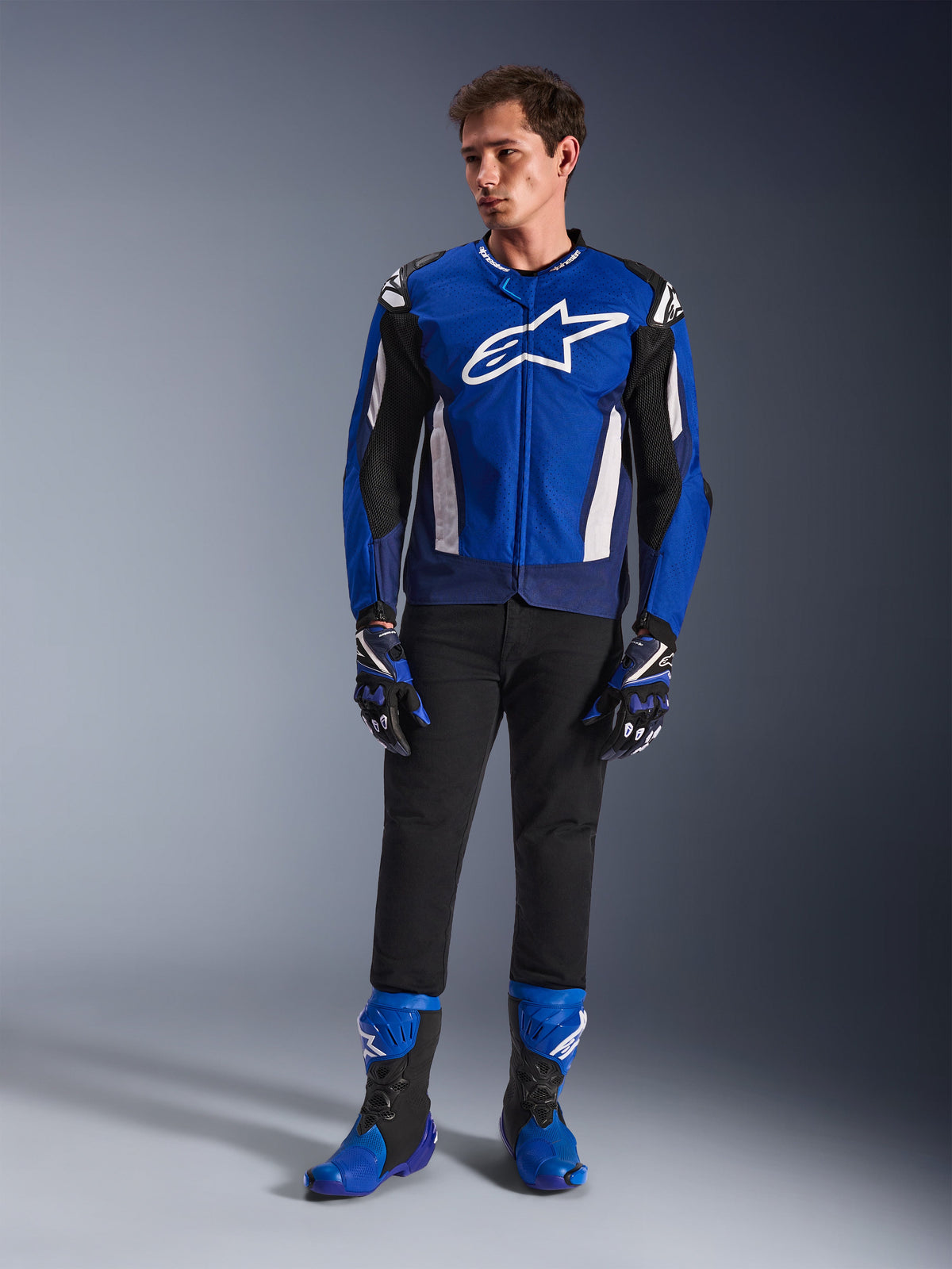 Modello che indossa la giacca Alpinestars T-GP Air in Bright Blue Navy White, abbinata a pantaloni neri, guanti da corsa coordinati blu e navy e stivali da moto blu e neri, scatto in studio che mostra un outfit completo da guida sportiva.