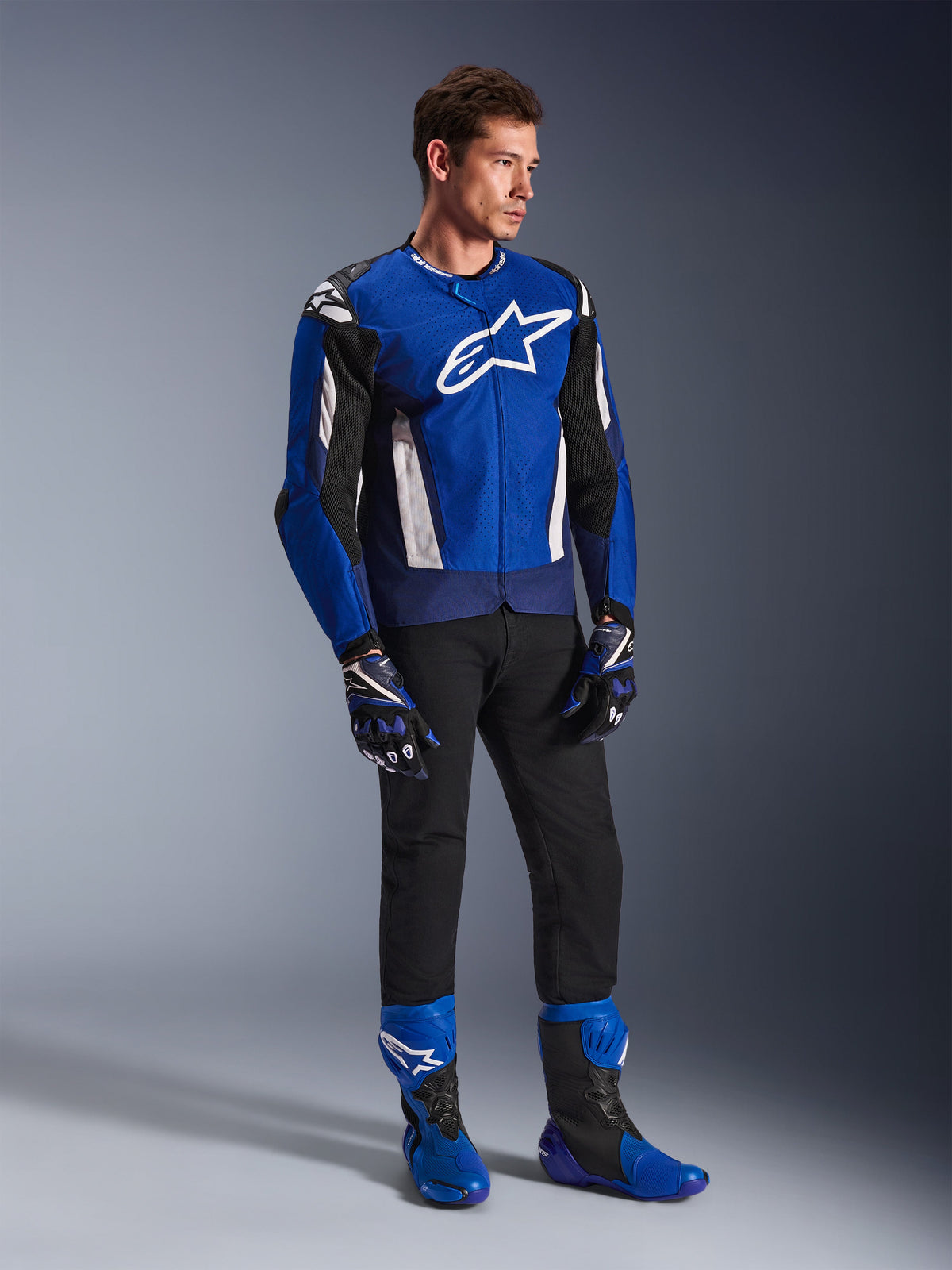 Alpinestars T-GP Air Jacket, giacca da moto sportiva in tessuto, blu brillante, navy e bianco, indossata con guanti blu e neri coordinati, pantaloni neri e stivali da corsa blu, con logo bianco sul petto e pannelli in mesh tecnico, scatto in studio a figura intera