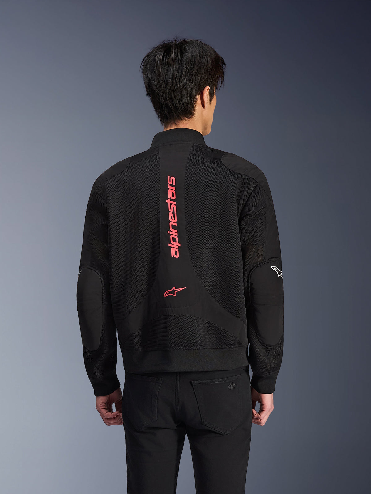 Vista posteriore di un pilota che indossa la giacca Alpinestars Flight Air in Black Bright Red, giacca da moto urbana stile bomber con costruzione in mesh 3D, con grande marchio Alpinestars verticale rosso e logo a stella sulla schiena, abbigliamento tecnico d'ispirazione street