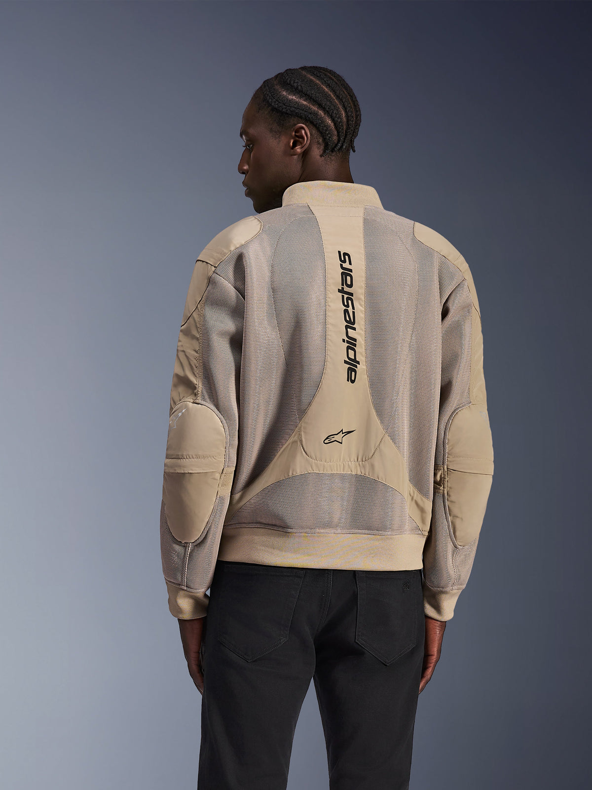 Giacca Alpinestars Flight Air, giacca da moto urbana, color sabbia, vista posteriore con ampi pannelli in mesh 3D e branding Alpinestars verticale sulla schiena, design ispirato ai bomber con orlo a costine, indossata da un modello con pantaloni neri