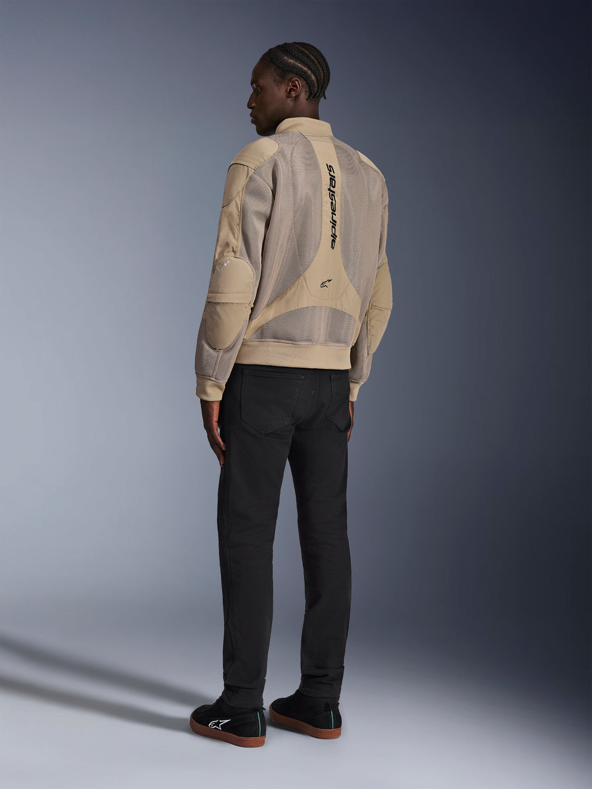Giacca Alpinestars Flight Air, giacca da moto urbana in tessuto, marrone, vista posteriore che mostra ampi pannelli in mesh 3D e logo verticale, silhouette stile bomber con colletto e orlo a costine, abbinata a pantaloni neri e scarpe nere per uno stile di guida di ispirazione street.