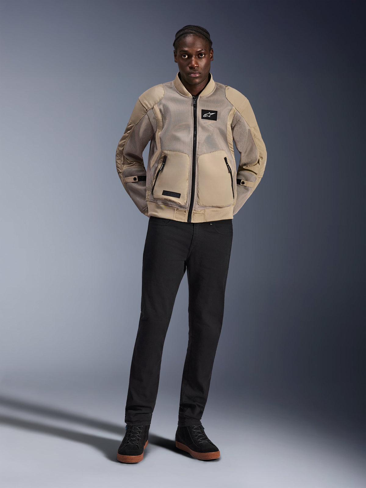 Modello che indossa la giacca Alpinestars Flight Air in marrone, giacca da moto urbana con ampi pannelli in mesh 3D, colletto e polsini a costine stile bomber, abbinata a pantaloni neri e scarpe da moto nere, abbigliamento contemporaneo street-ready