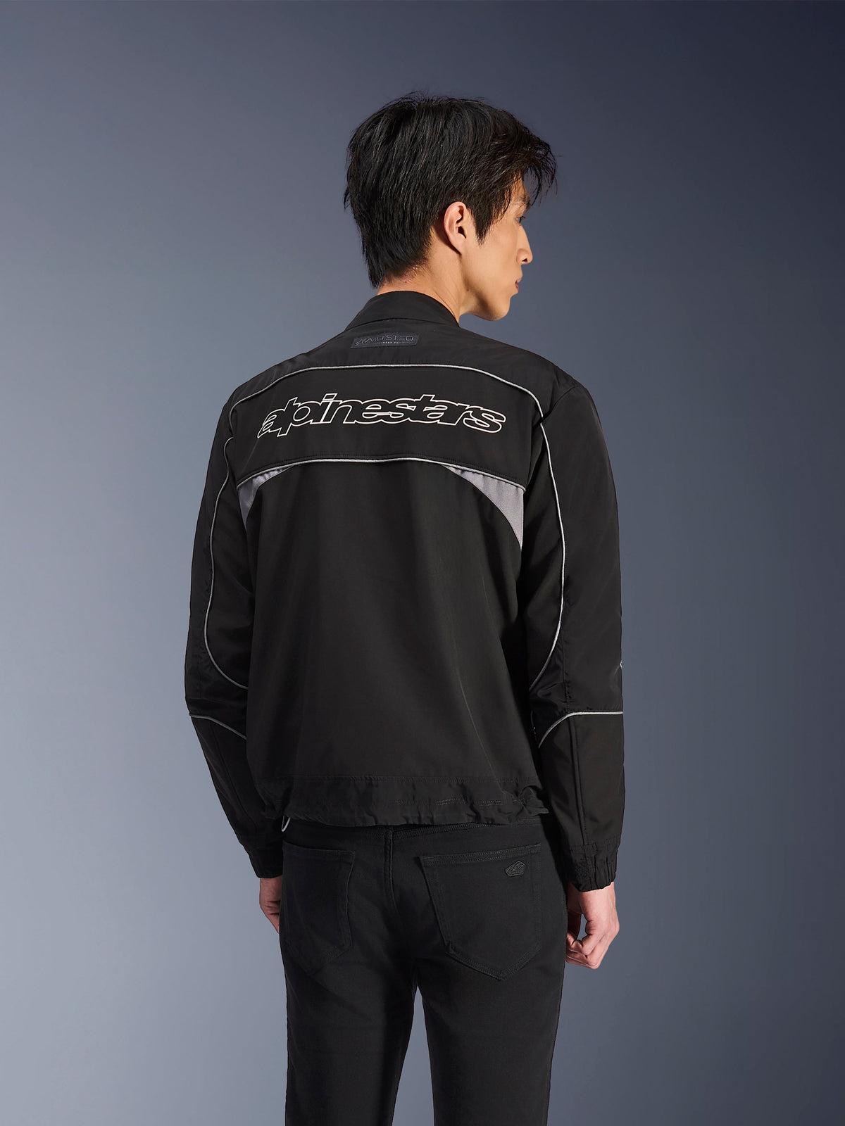 Giacca Alpinestars Unite, giacca da moto urbana, nero con accenti grigio scuro, vista posteriore con scritta Alpinestars bianca sulle spalle, profili e pannelli riflettenti, design streetwear in softshell minimalista