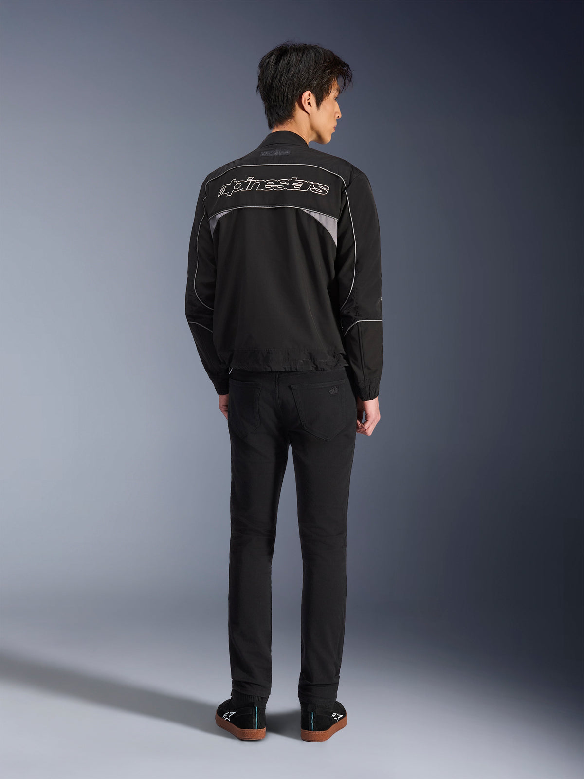 Giacca Alpinestars Unite, giacca da moto nero e grigio scuro, vista posteriore con logo Alpinestars profilato sulla schiena, pannelli riflettenti grigi, design urbano minimalista, indossata con pantaloni neri e scarpe nere