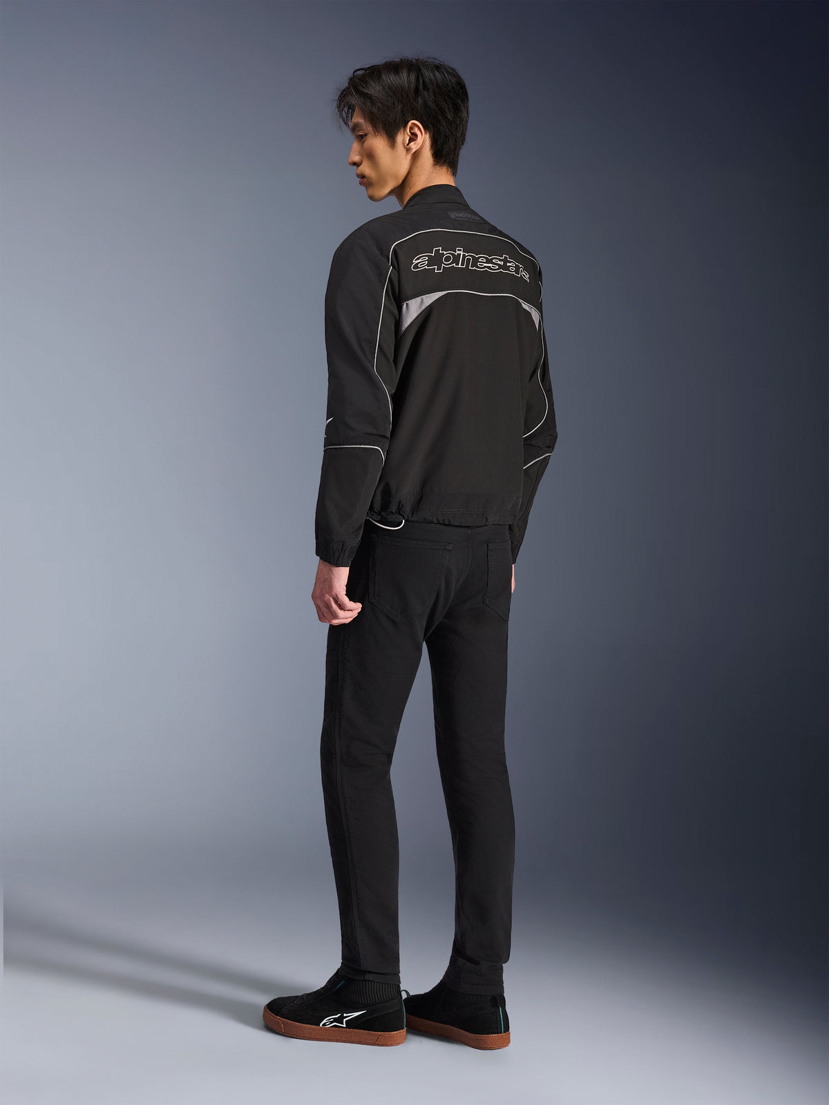 Alpinestars Unite Jacket, giacca da moto urban, nero e grigio scuro, vista posteriore con grande logo Alpinestars profilato, profili e pannelli riflettenti, design softshell minimalista, indossata con pantaloni neri e calzature casual