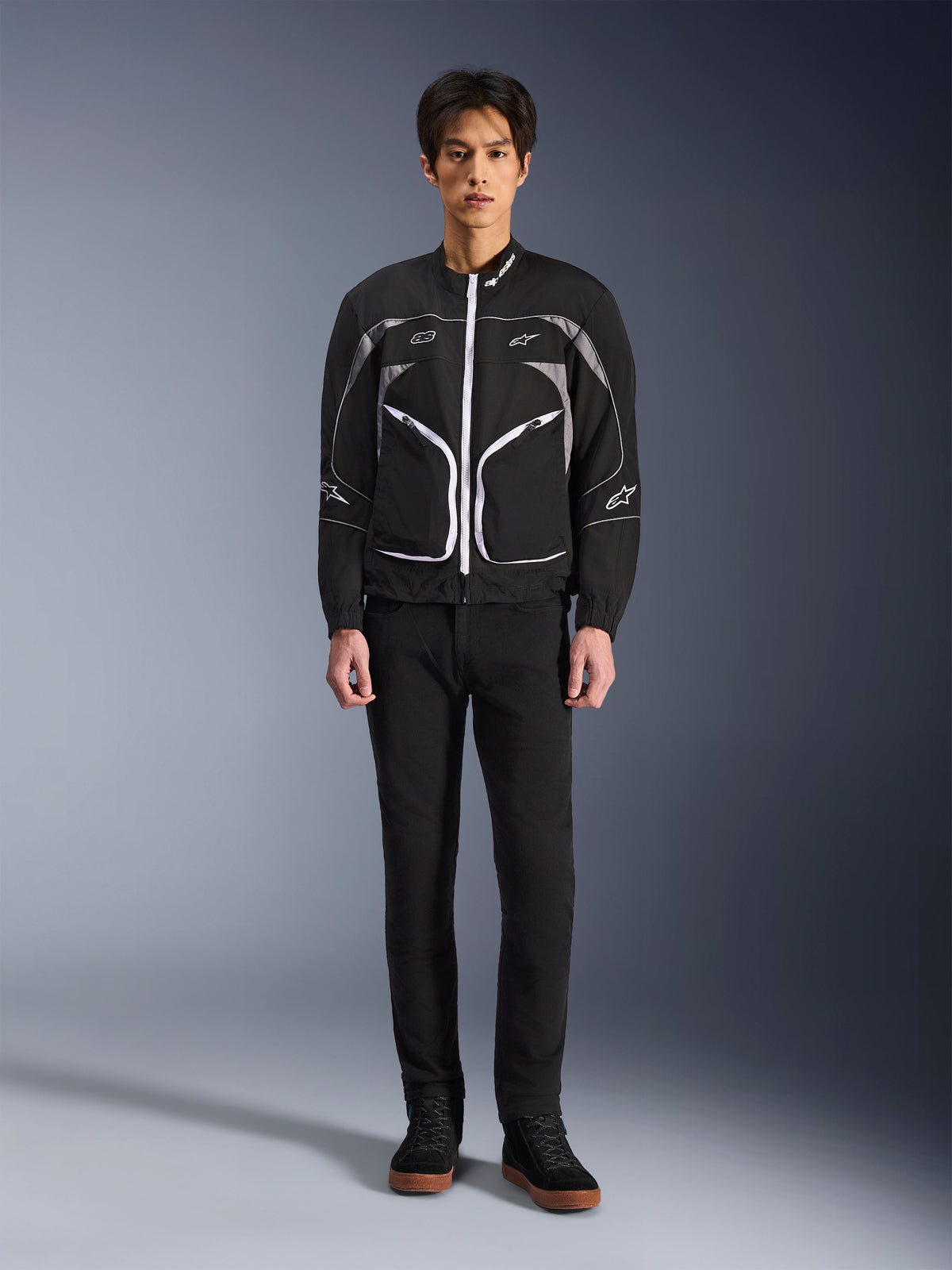 Giacca Alpinestars Unite, giacca da moto, nero e grigio scuro, indossata da un modello con pantaloni neri e stivali neri, con cerniere bianche a contrasto, profili riflettenti e design urbano minimalista