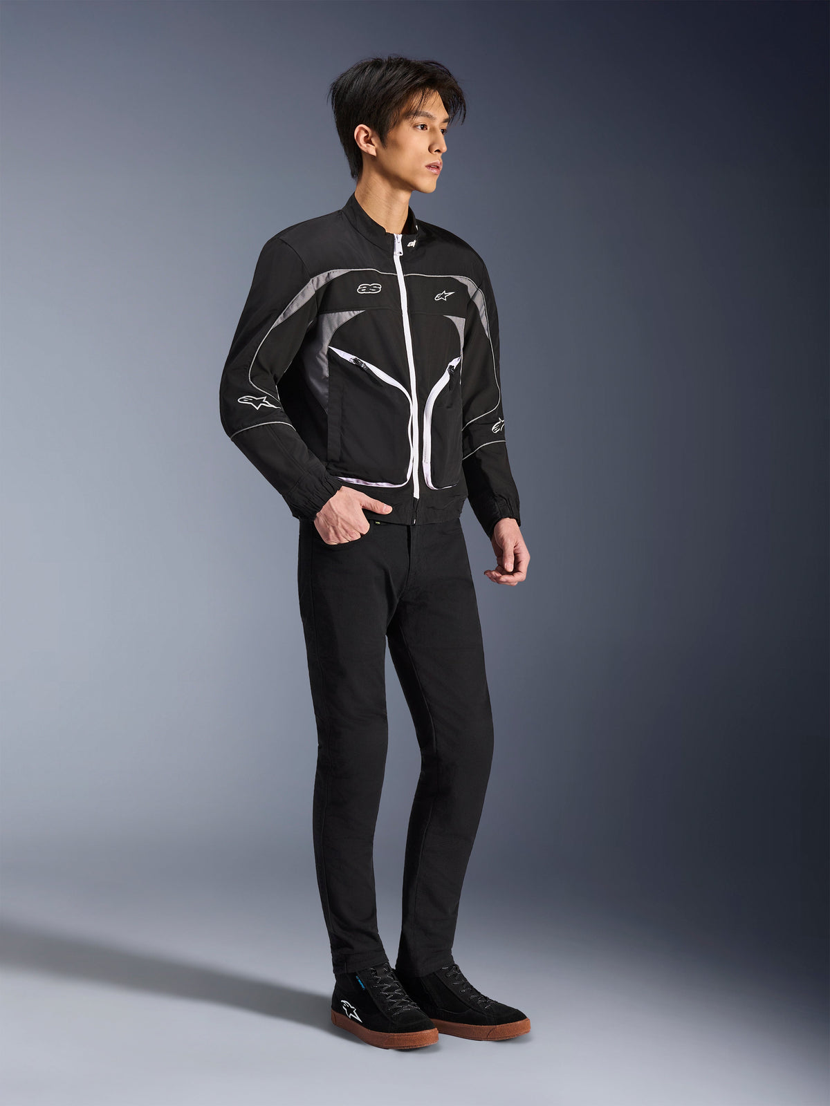 Modello che indossa la giacca Alpinestars Unite in nero e grigio scuro, giacca da moto urbana con cerniere bianche e dettagli riflettenti, abbinata a pantaloni neri slim-fit e sneakers, abbigliamento da moto minimalista d'ispirazione street