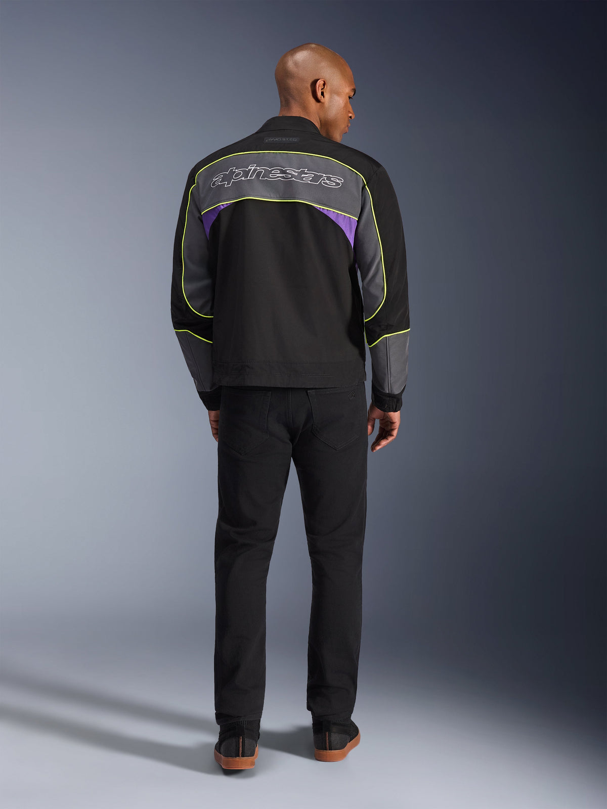 Alpinestars Unite Jacket, giacca da moto urbana in Black Asphalt Violet, vista posteriore che mostra pannelli grigi con accenti viola e profili giallo neon, logo Alpinestars a scritta sulle spalle, indossata con pantaloni neri