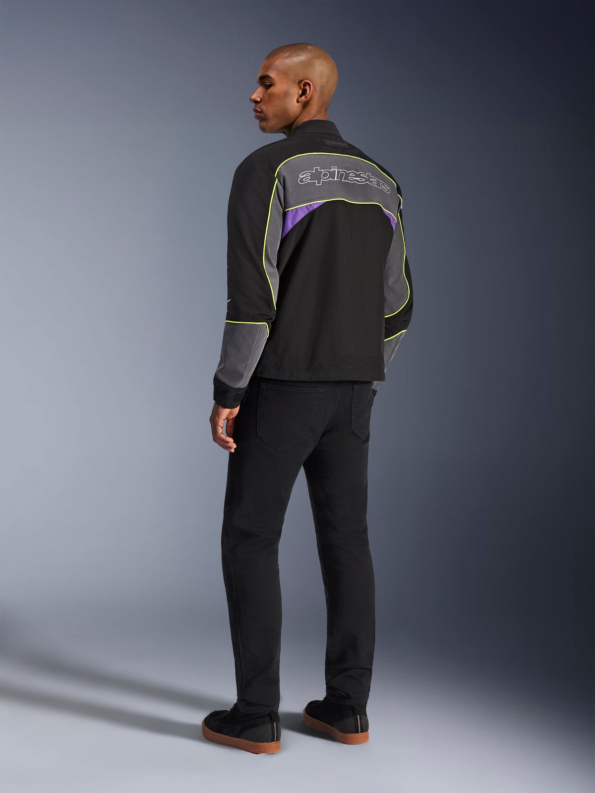 Alpinestars Unite Jacket, giacca da moto urbana, Black Asphalt Violet, vista posteriore di tre quarti su modello, softshell nero con pannelli grigi e accenti viola, profili riflettenti giallo neon, logo Alpinestars sulla parte superiore della schiena, abbinata a pantaloni neri e scarpe casual nere