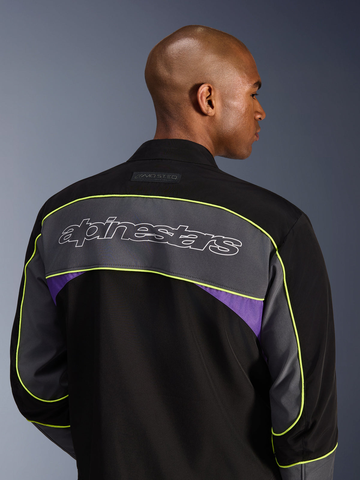 Giacca Alpinestars Unite, giacca da moto urban, Black Asphalt Violet, vista posteriore che mostra un ampio pannello grigio sulle spalle con logo Alpinestars, bordature giallo neon e accenti viola su una costruzione in softshell a 3 strati