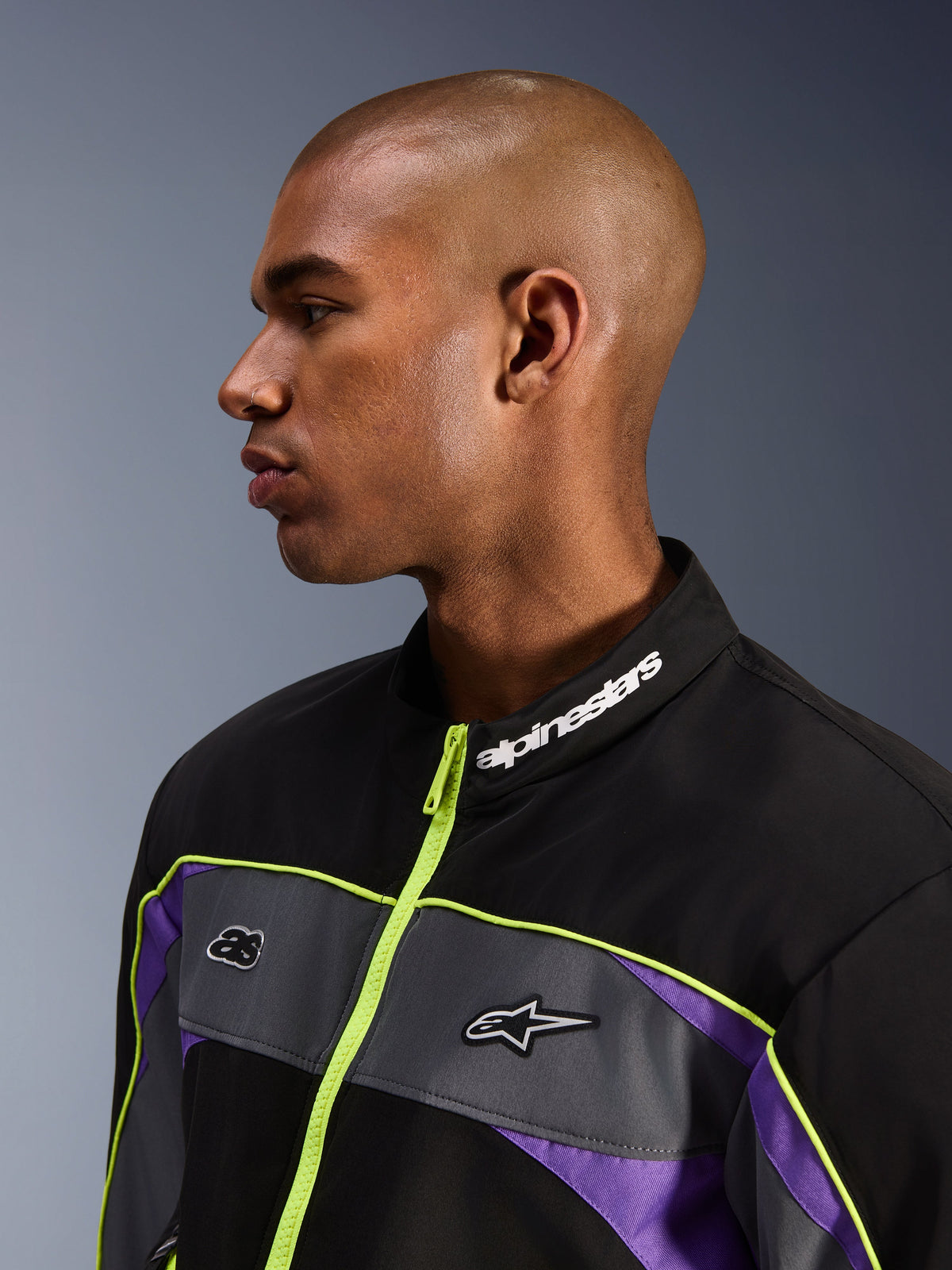 Giacca Alpinestars Unite, Black Asphalt Violet, vista di profilo di un modello con focus sul colletto alto con branding Alpinestars bianco, cerniera giallo neon, pannelli grigi sul petto con loghi, accenti viola, stile streetwear motociclistico urbano minimalista