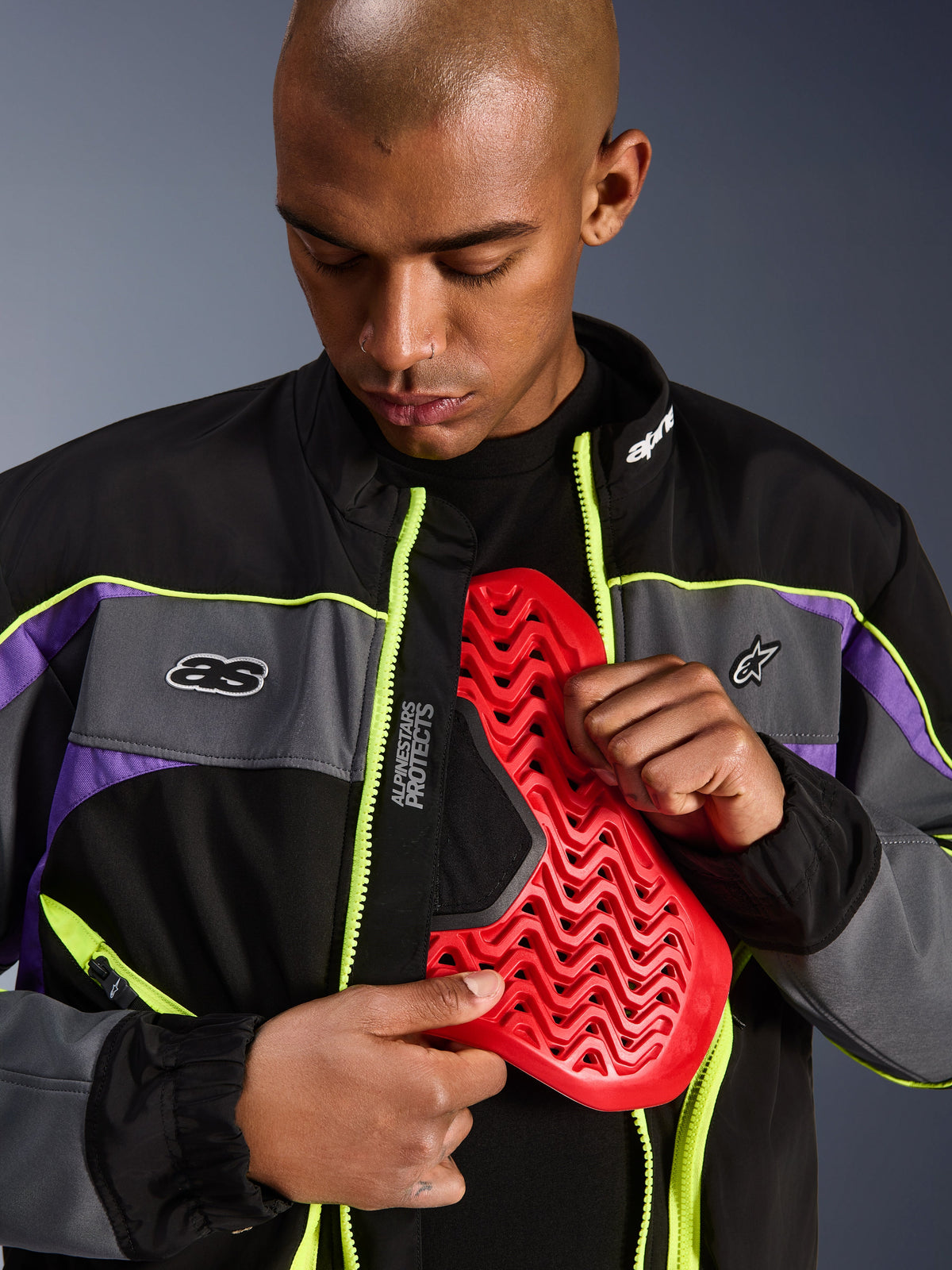 Giacca Alpinestars Unite, giacca da moto urbana, nero asfalto e viola con accenti giallo neon, modello maschile che mostra un inserto per protezione toracica Nucleon rosso all'interno della giacca aperta, estetica streetwear minimalista per guida urbana