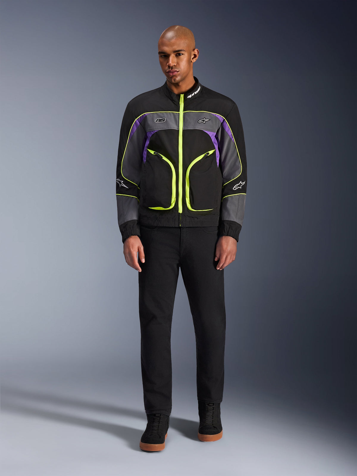 Modello che indossa la giacca Alpinestars Unite in Black Asphalt Violet, caratterizzata da una base nera con pannelli grigi e viola e vivaci cerniere e finiture giallo neon, abbinata a pantaloni e scarpe neri, giacca da moto urbana minimalista.