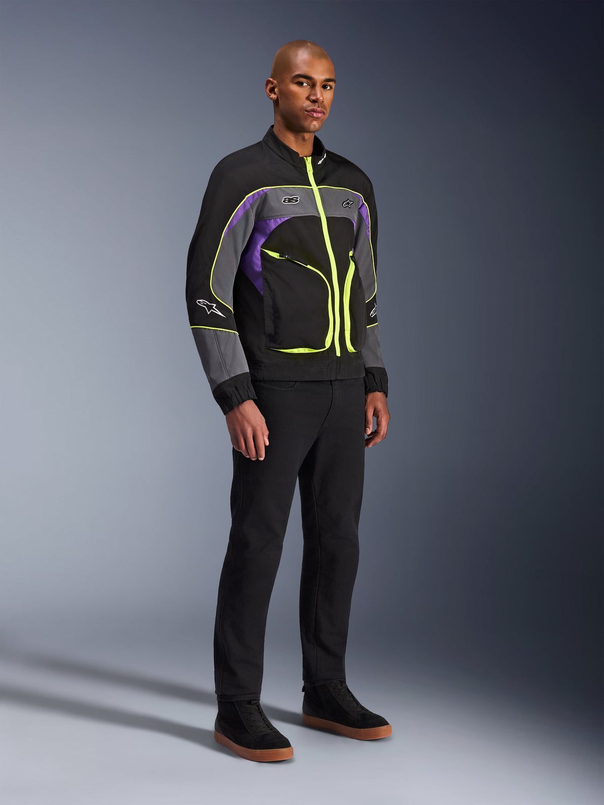 Giacca Unite Alpinestars, giacca da moto urbana, Black Asphalt Violet con accenti giallo neon, indossata da un modello con pantaloni neri e sneakers, stile streetwear contemporaneo per la guida urbana