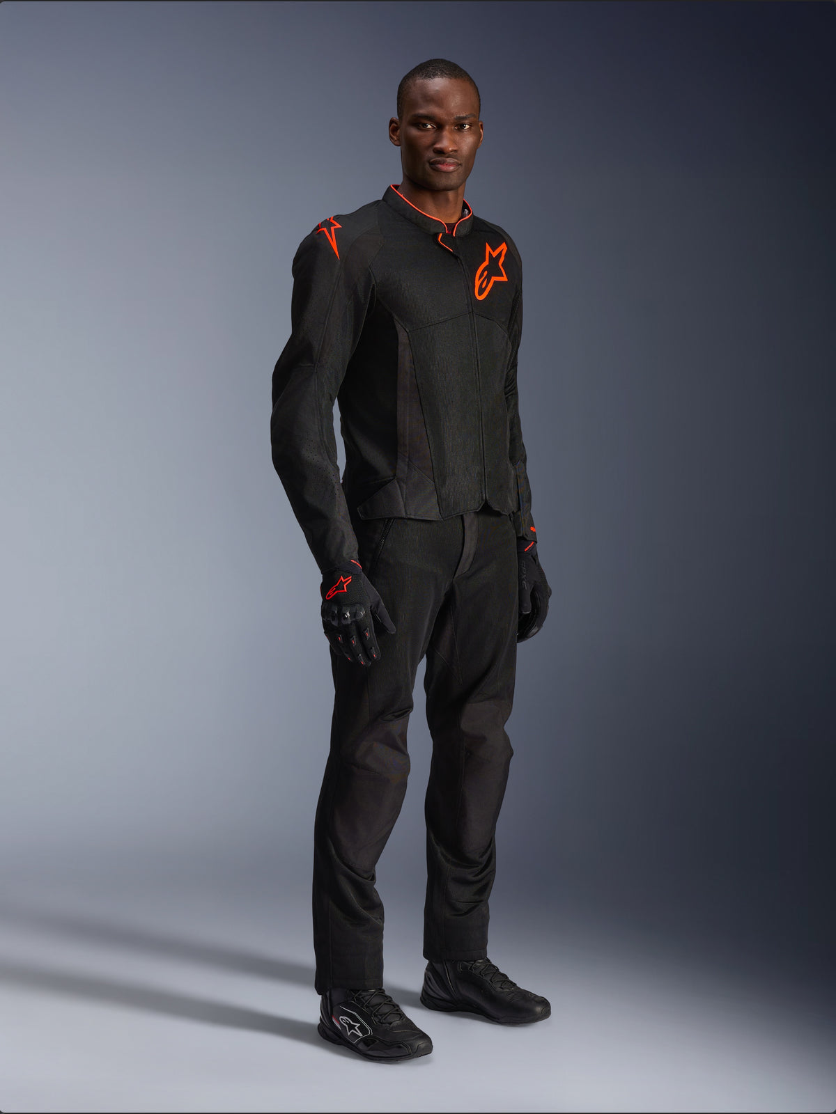 Modello che indossa la giacca Alpinestars Viper Air V4 in nero e rosso fluo, con pannelli in rete e loghi a stella rossi sul petto e sulla spalla, abbinata a pantaloni da moto, guanti e scarpe da guida neri coordinati, completo professionale per la guida su strada