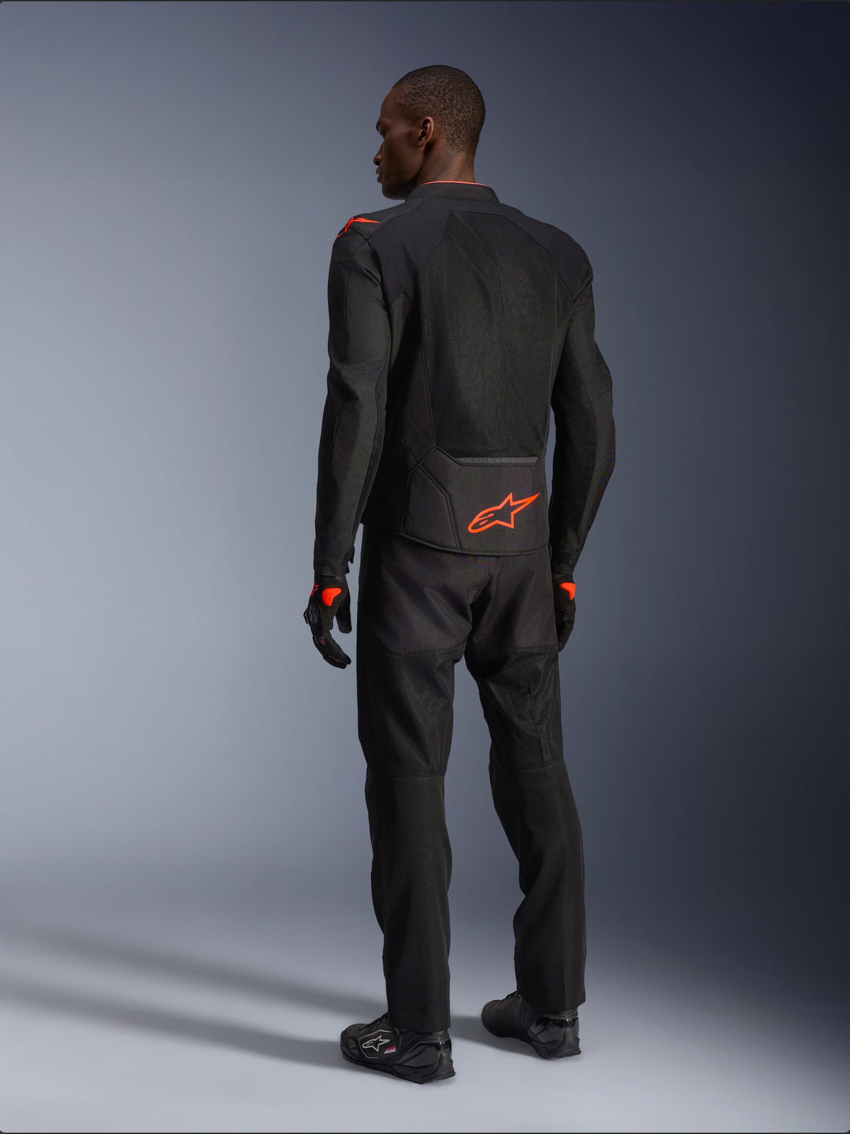 Pilota uomo visto da dietro che indossa la giacca Alpinestars Viper Air V4, nero e rosso fluo, con logo Astars arancione sulla parte bassa della schiena e sulle spalle, costruzione in tessuto mesh, abbinata a pantaloni e stivali da moto neri coordinati.