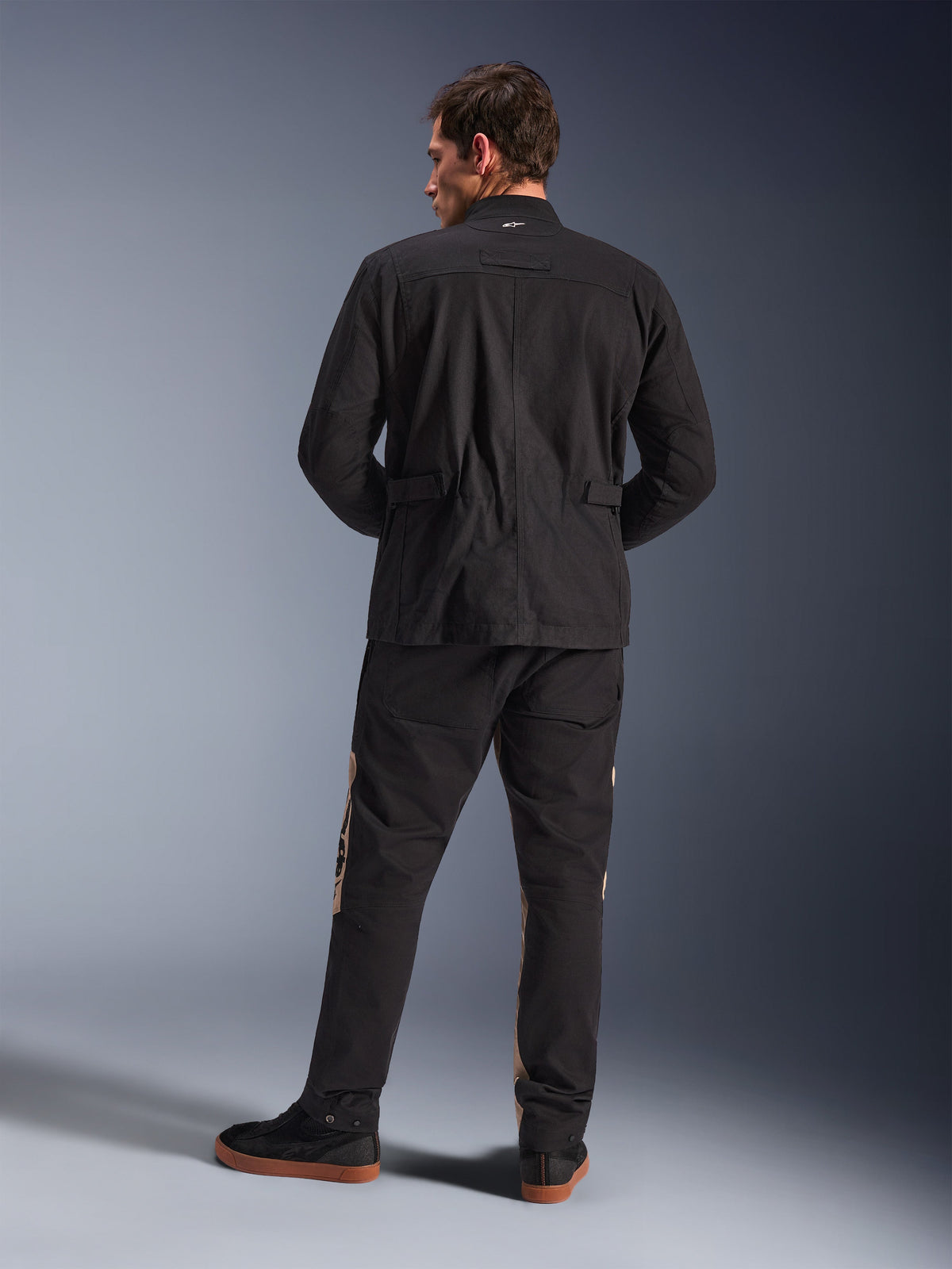 Vista posteriore della Giacca Alpinestars Dice in nero, giacca da moto urban stile field con spalle plissettate e regolatori in vita, abbinata a pantaloni da moto neri e marrone chiaro e sneakers, stile contemporaneo per gli spostamenti urbani