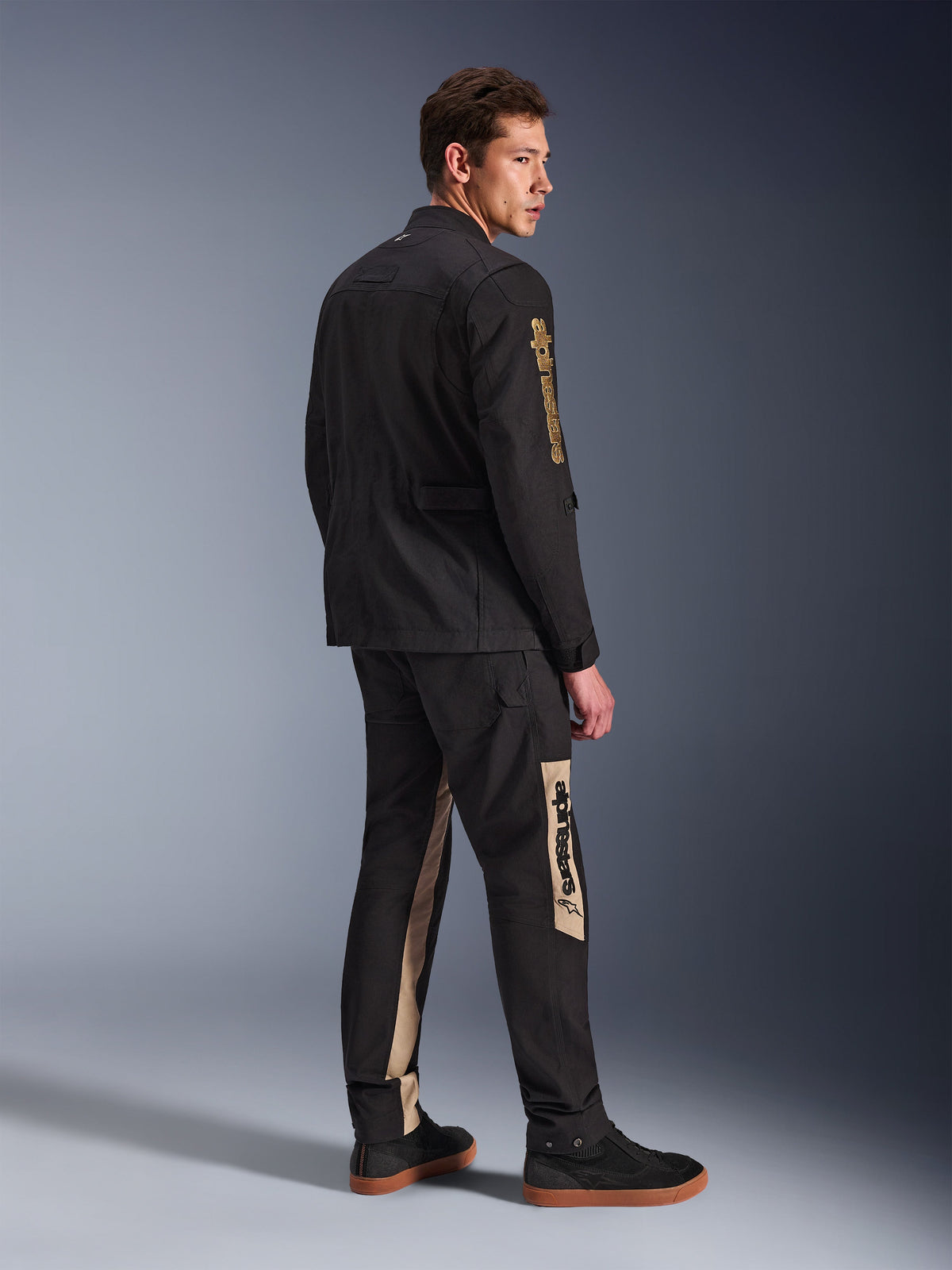 Giacca Alpinestars Dice, giacca da moto urbana, nero, vista posteriore a 3/4 su modello, caratterizzata da un'estetica vintage field jacket con logo Alpinestars dorato sulla manica, indossata con pantaloni da moto coordinati nero e beige e sneakers nere