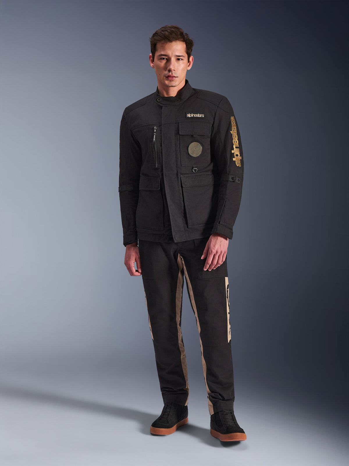 Modello che indossa la giacca Alpinestars Dice Jacket, giacca da moto urbana nera con stile vintage da field jacket, quattro tasche anteriori funzionali, logo Alpinestars sulla manica e sul petto, abbinata a pantaloni neri e beige, abbigliamento moderno per la guida urbana
