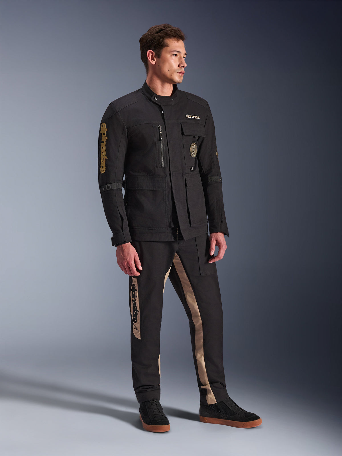 Modello che indossa la giacca Alpinestars Dice Jacket in nero, giacca da moto urbana dall'estetica vintage field jacket, con tasche utility multiple e logo dorato sulla manica, abbinata a pantaloni coordinati neri e tan e sneaker nere.