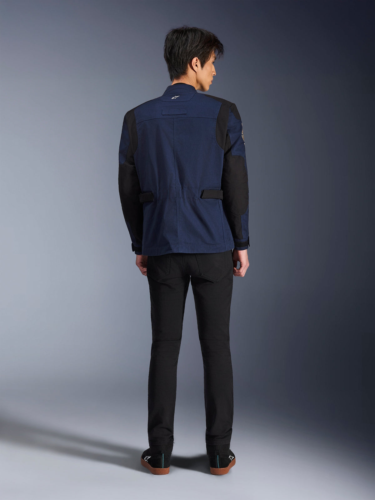 Modello che indossa la Giacca Dice Alpinestars in Blu Scuro e Nero, vista posteriore, giacca da moto urban stile field jacket con spalle plissettate e regolatori in vita, abbinata a pantaloni neri e sneakers nere.