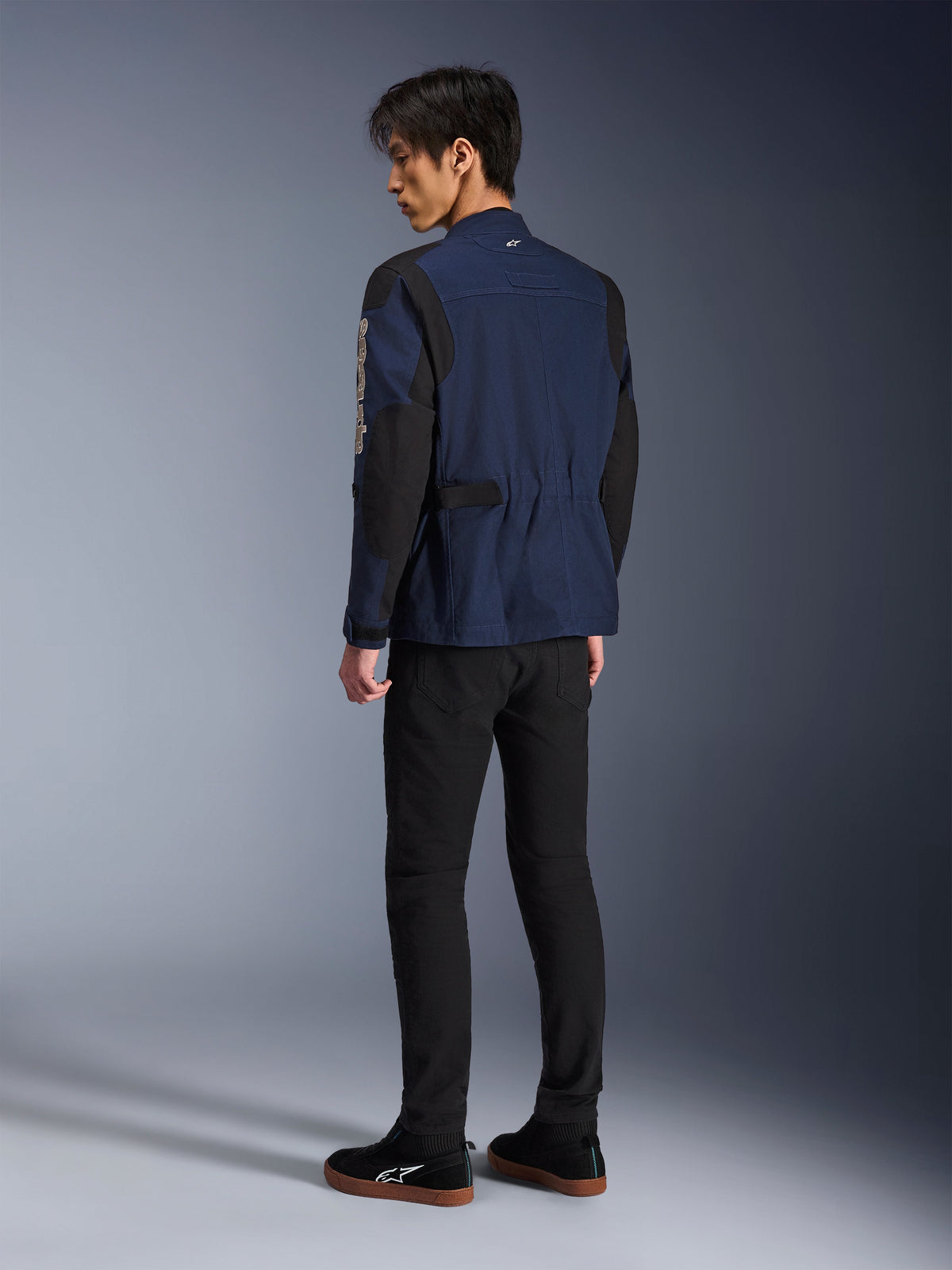 Modello uomo visto di spalle che indossa la giacca Alpinestars Dice in blu scuro e nero, abbinata a pantaloni neri e sneaker nere, giacca da moto urbana stile field jacket con spalle plissettate e linguette di regolazione in vita.