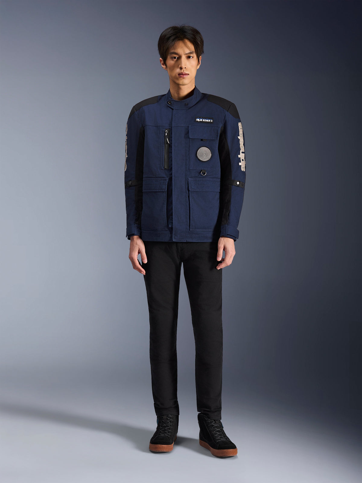 Alpinestars Dice Jacket, giacca da moto urban, blu scuro e nero, indossata da un modello con pantaloni neri e scarpe nere, design moderno da giacca da campo con tasche multiple, cerniera verticale sul petto e marchio Alpinestars bianco sulle maniche.