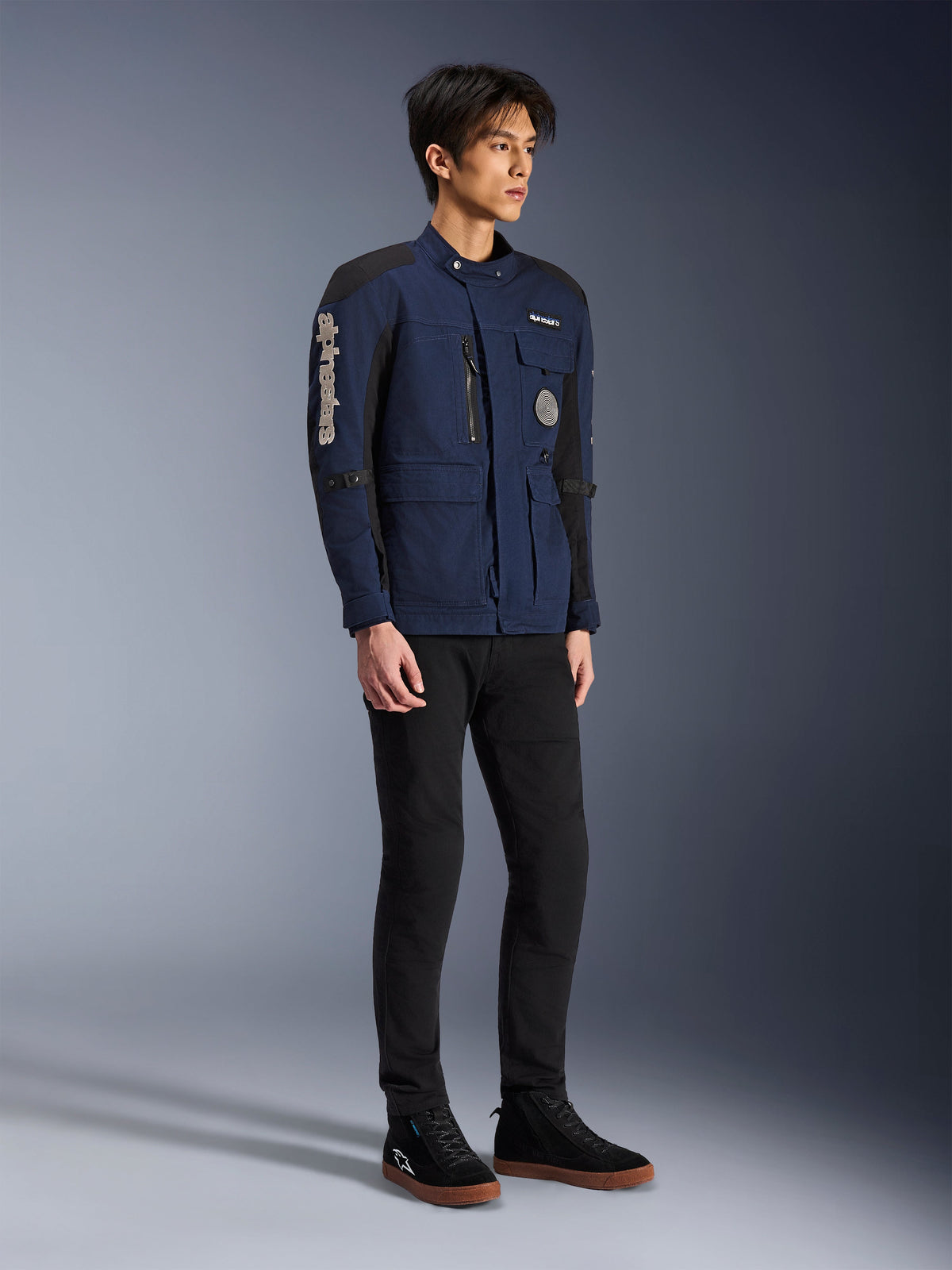 Giacca Alpinestars Dice, blu scuro e nero, moderna giacca da moto in stile field indossata da un modello con pantaloni neri e sneakers alte nere, dotata di molteplici tasche utility, marchio Alpinestars verticale sulla manica e toppe in tessuto, abbigliamento tecnico urbano contemporaneo