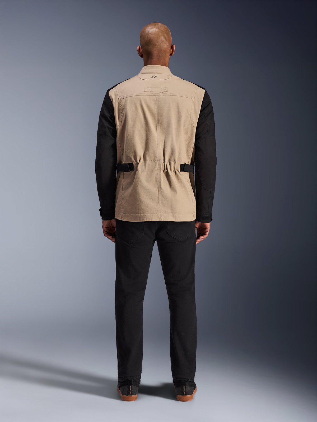 Giacca Alpinestars Dice, giacca da moto urbana, color sabbia e nero, vista posteriore su modello, caratterizzata da una struttura in twill di policotone, cinturini regolabili in vita e piccolo logo ricamato sul colletto, indossata con pantaloni neri