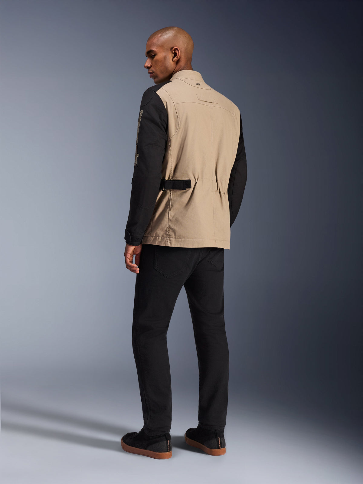 Giacca Alpinestars Dice, giacca da moto urbana, Aluminum Black con busto tan e maniche nere, vista posteriore su modello, design robusto ispirato al workwear con regolatori in vita, abbinata a pantaloni da moto neri.