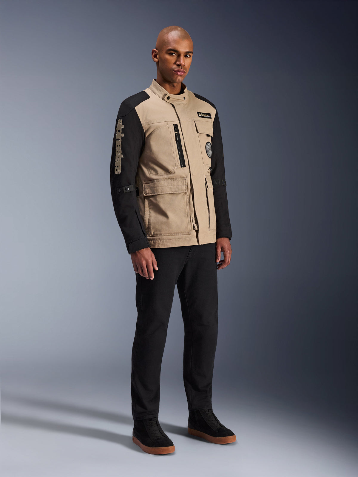 Alpinestars Giacca Dice, giacca da moto urbana, Aluminum Black e tan, design bicolore con maniche nere e busto khaki, indossata da un modello con pantaloni neri e sneakers nere, estetica ispirata al workwear con tasche utility e marchio ricamato sulla manica.