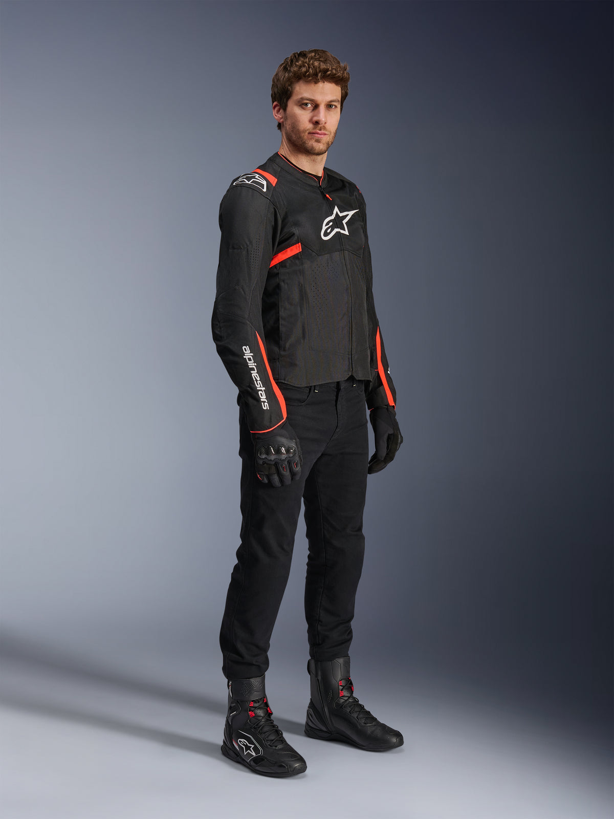 Modello che indossa la giacca Alpinestars T-Sps Air V2, nera con accenti rosso fluo, abbinata a pantaloni da moto neri, guanti e stivali neri, mettendo in risalto il design sportivo con pannelli in rete e loghi Alpinestars bianchi.
