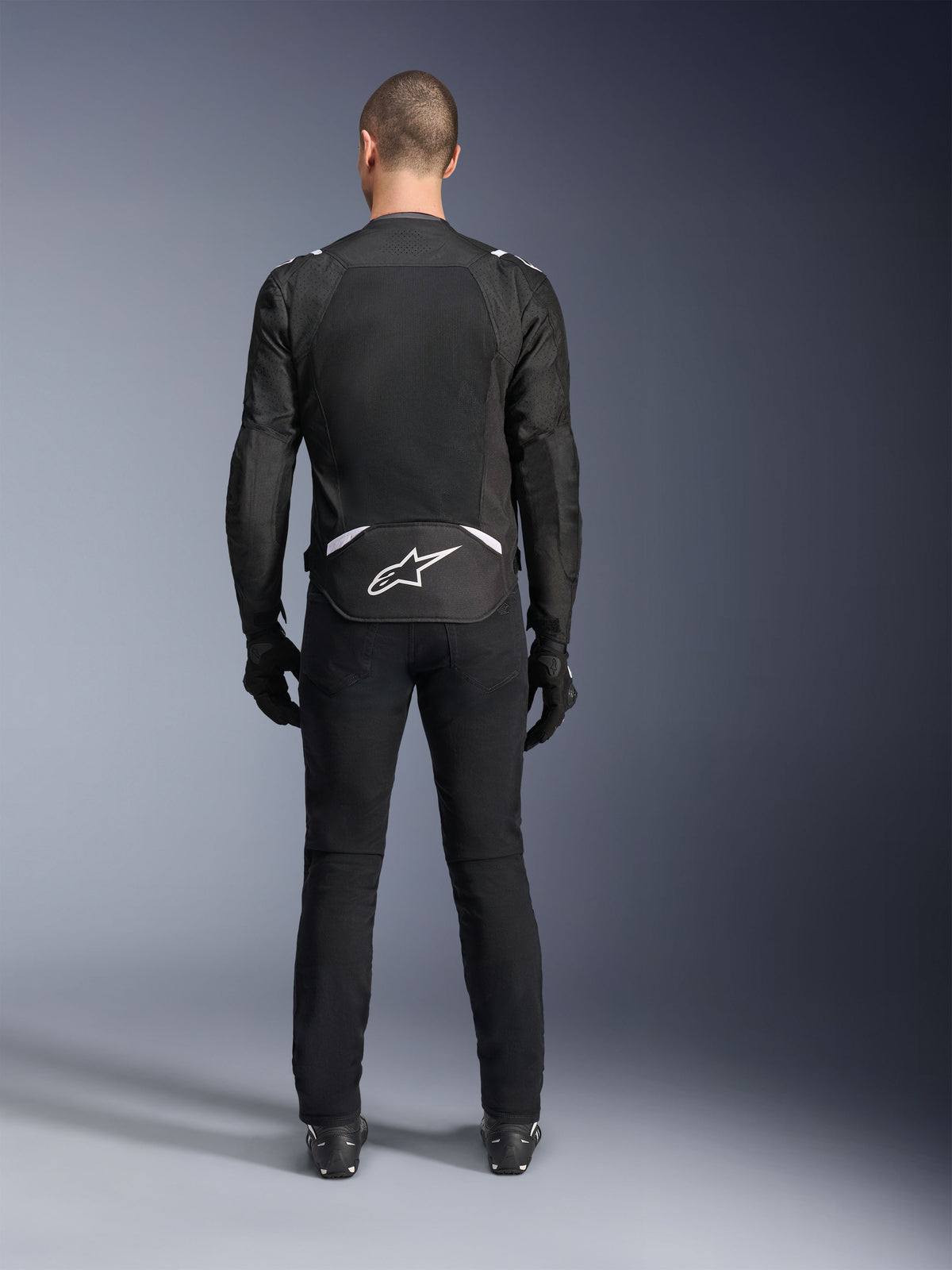 Giacca Alpinestars T-Sps Air V2, vista posteriore, giacca da moto in tessuto nero e bianco, dotata di un ampio pannello posteriore in mesh per il massimo flusso d'aria e logo Astars bianco sulla parte bassa della schiena, indossata con pantaloni e guanti da moto neri