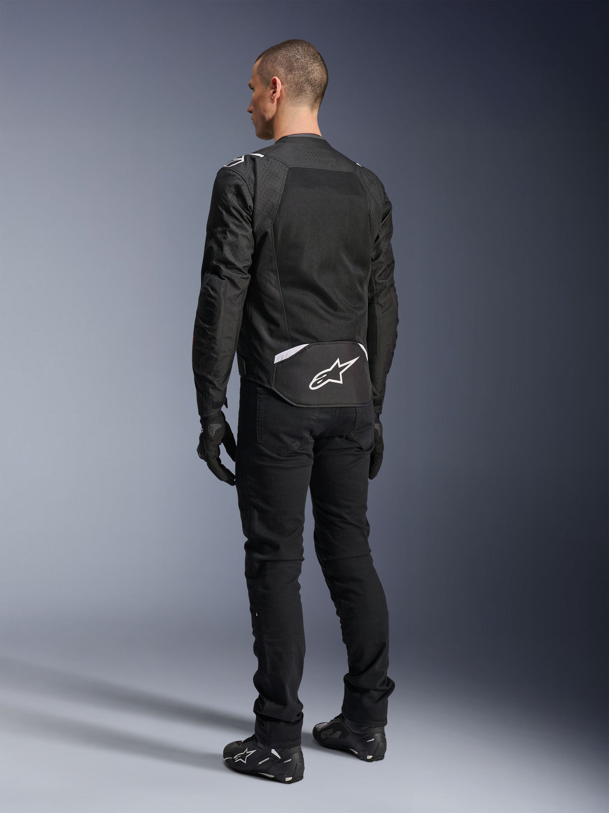 Giacca Alpinestars T-Sps Air V2, giacca da moto in tessuto, nera, vista posteriore con ampio pannello di ventilazione in mesh e logo Alpinestars bianco sulla parte bassa della schiena, abbinata a pantaloni e guanti neri per un look urbano sportivo.