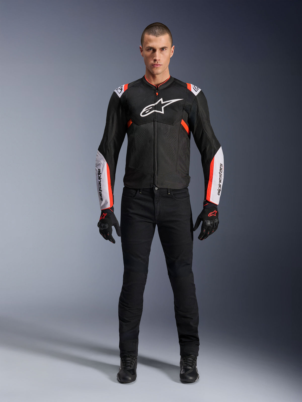 Modello che indossa la giacca Alpinestars T-Sps Air V2 in Black White Red Fluo, caratterizzata da ampi pannelli in mesh tecnico e logo bianco su petto e maniche, abbinata a pantaloni tecnici da guida e guanti neri, abbigliamento da moto sportivo urbano