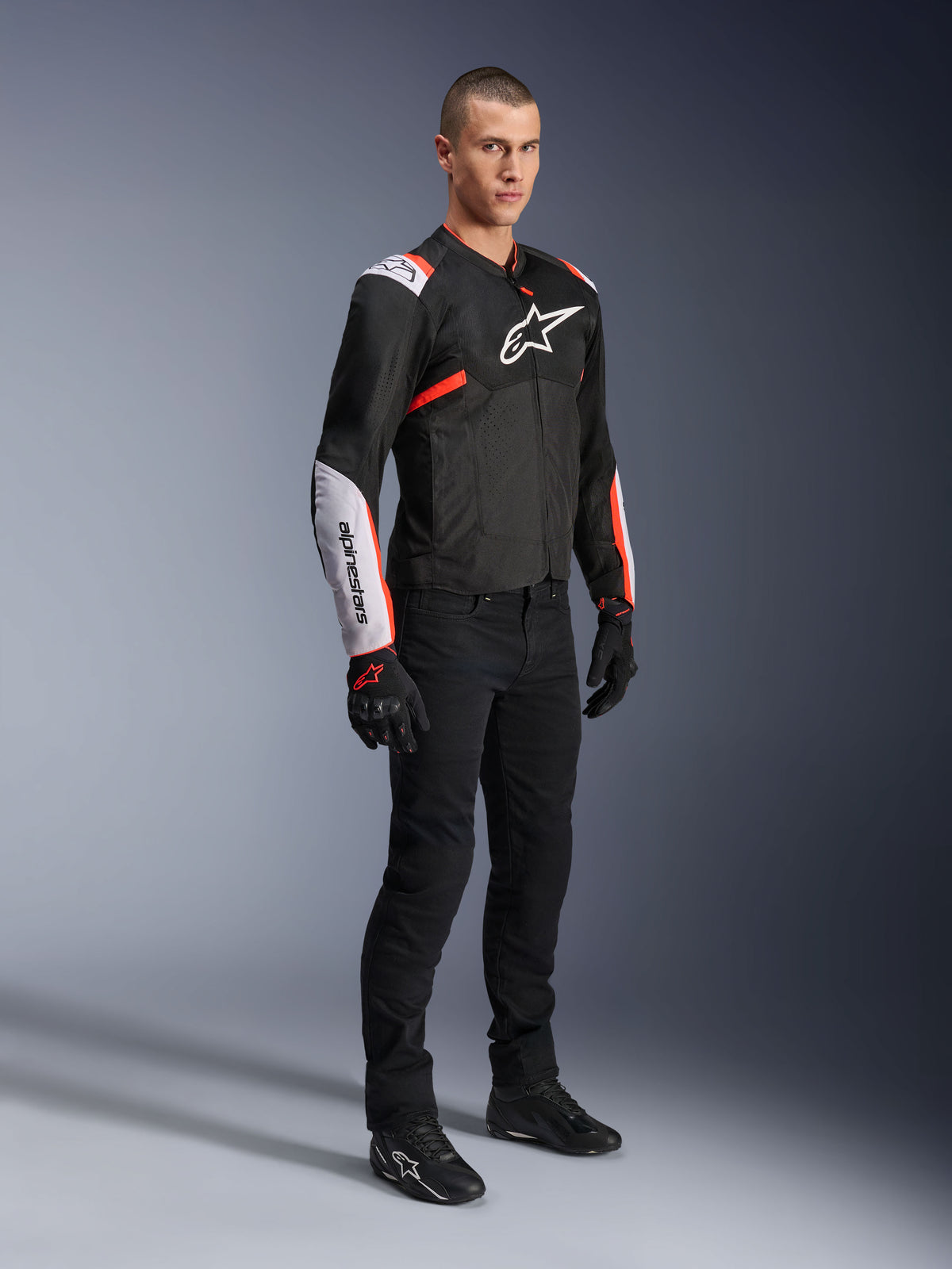 Giacca Alpinestars T-Sps Air V2, Nero Bianco Rosso Fluo, indossata da un modello uomo con pantaloni e guanti da moto neri, giacca da moto in tessuto con ampi pannelli in mesh, sezioni bianche su spalle e avambracci e logo Alpinestars sul petto.